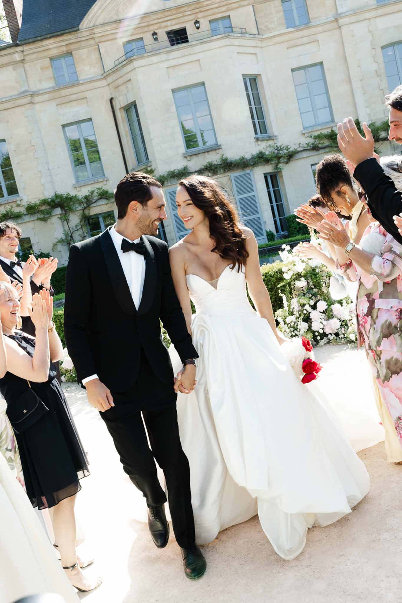 Couple walking guest line-up past white peony arrangements before vine-covered chateau with grey shutters