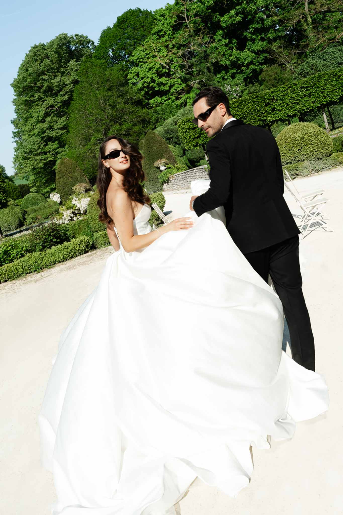 Bride in white ball gown and groom in black tuxedo walking through a formal French garden wearing sunglasses