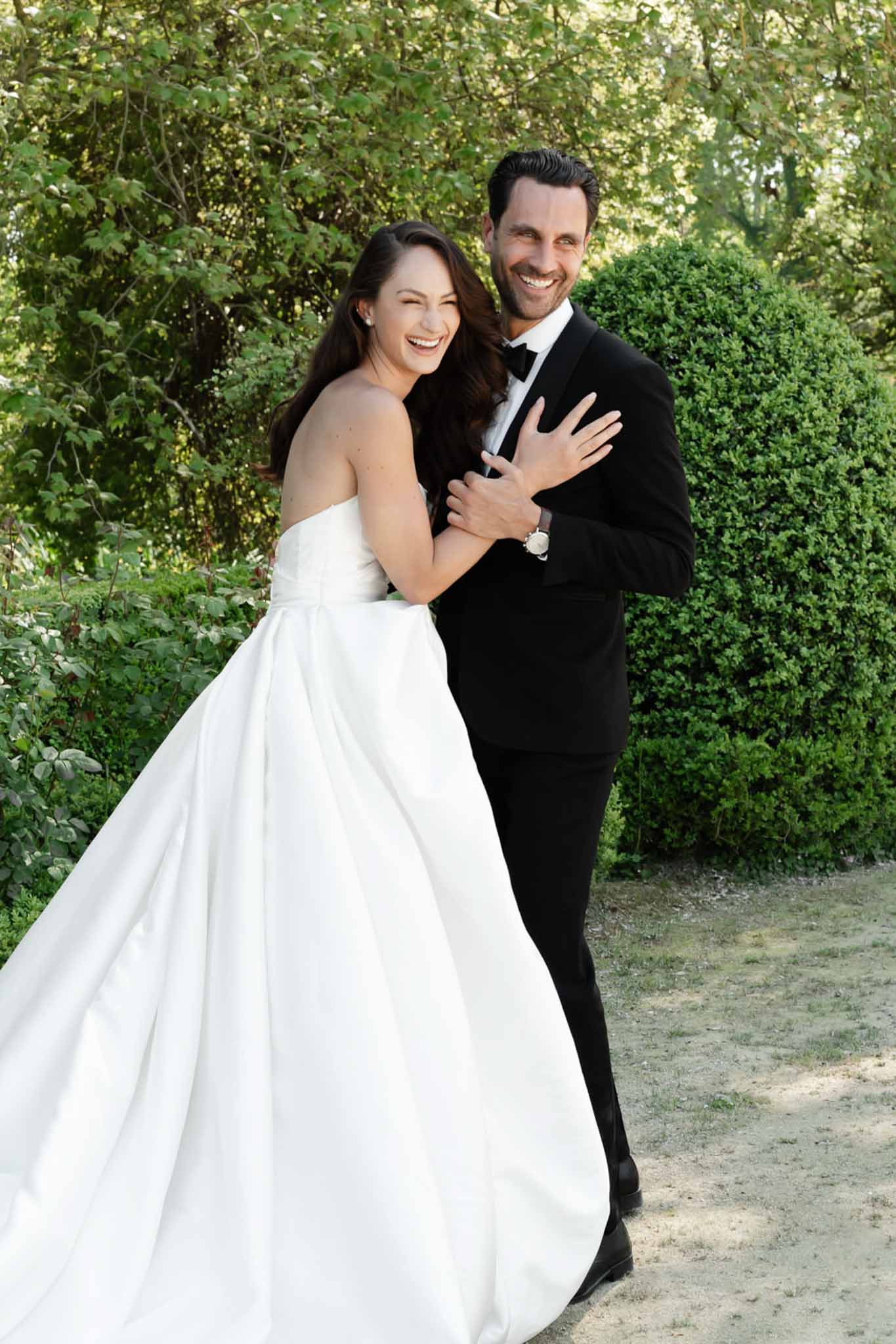 Laughing bride and groom embracing in a garden with boxwood hedges, bride in white strapless ballgown