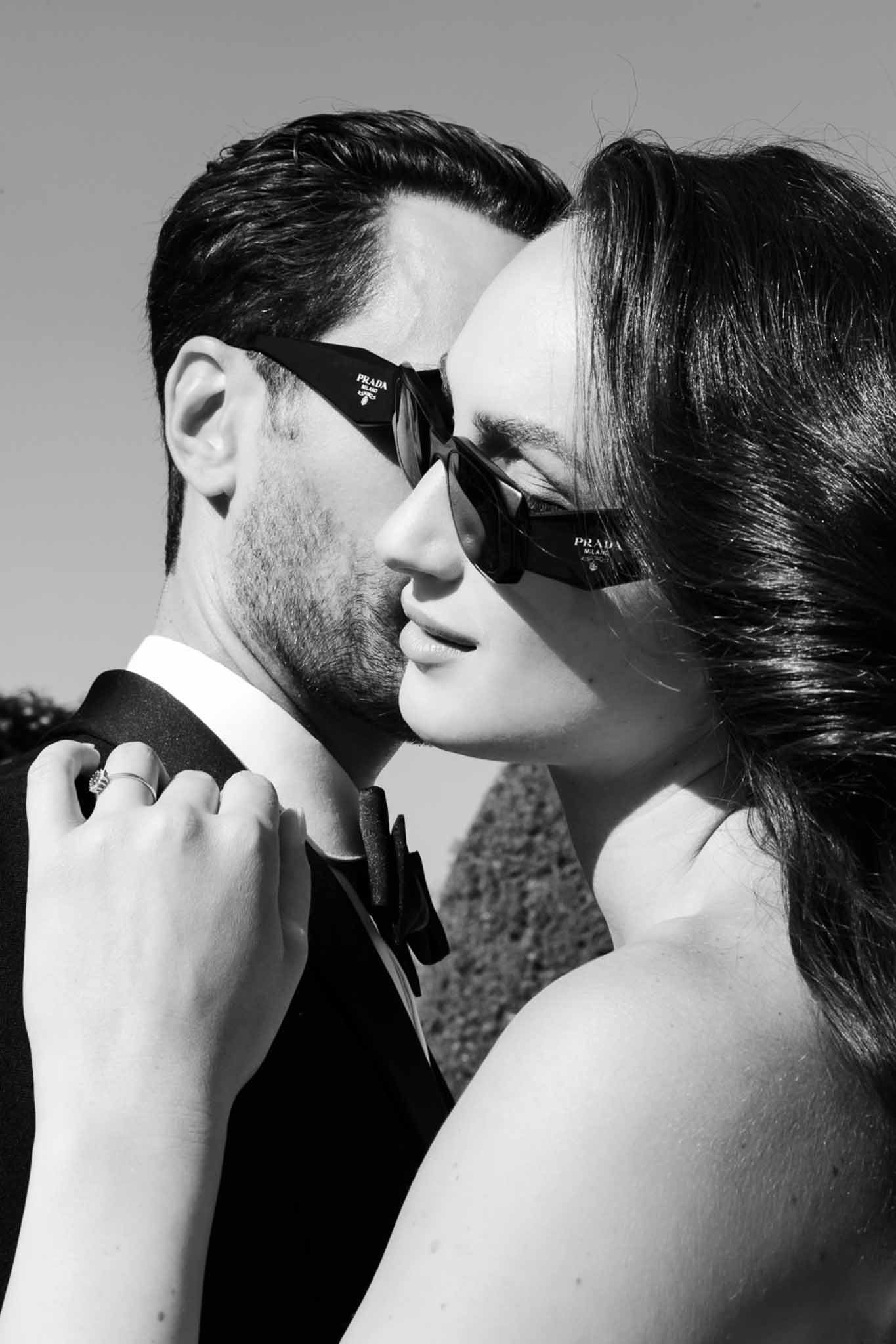 Bride and groom in matching Prada sunglasses nearly touching faces with diamond ring on lapel in B&W