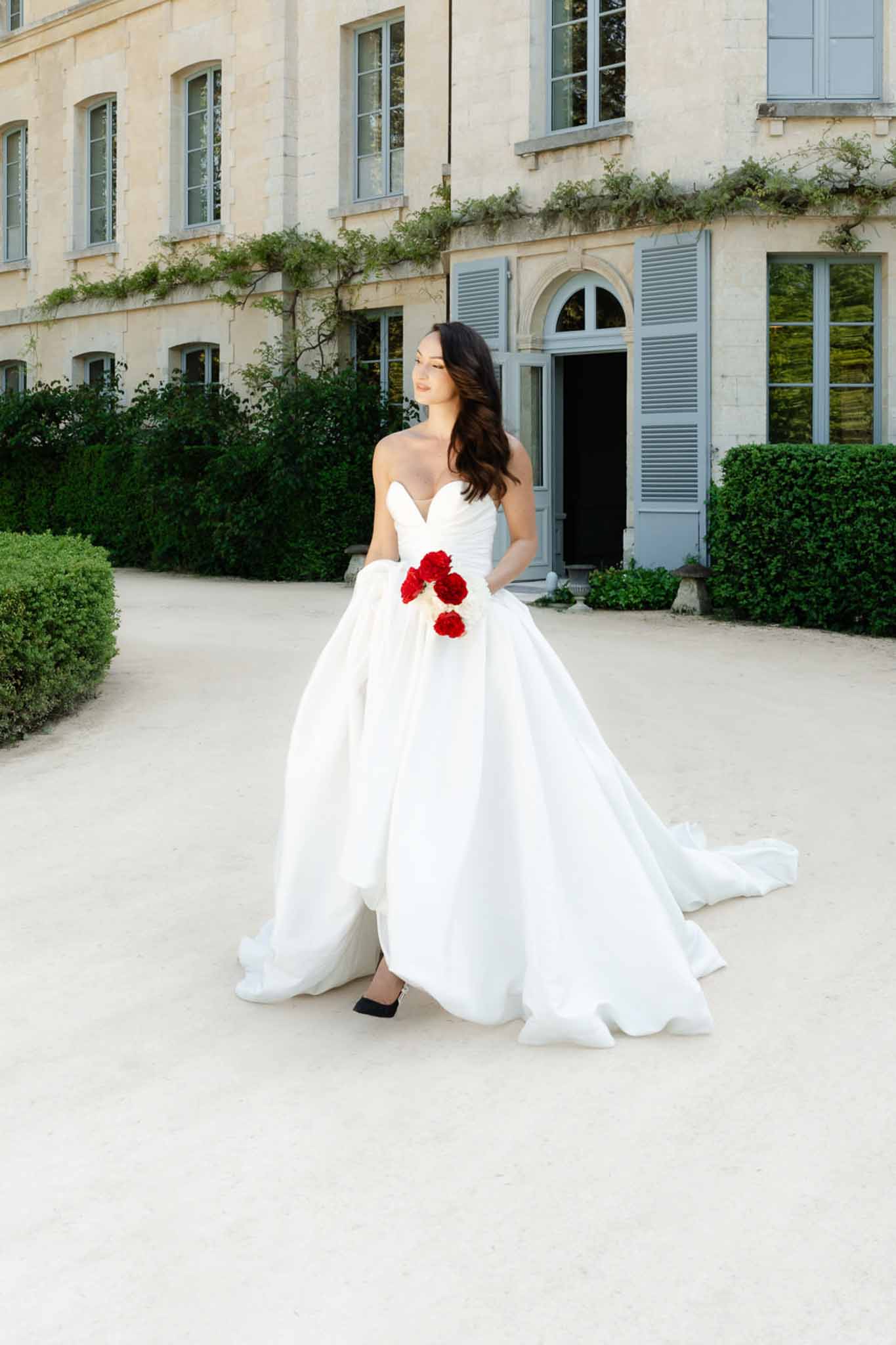 Bride in strapless ivory ballgown holding deep red rose bouquet on chateau gravel courtyard