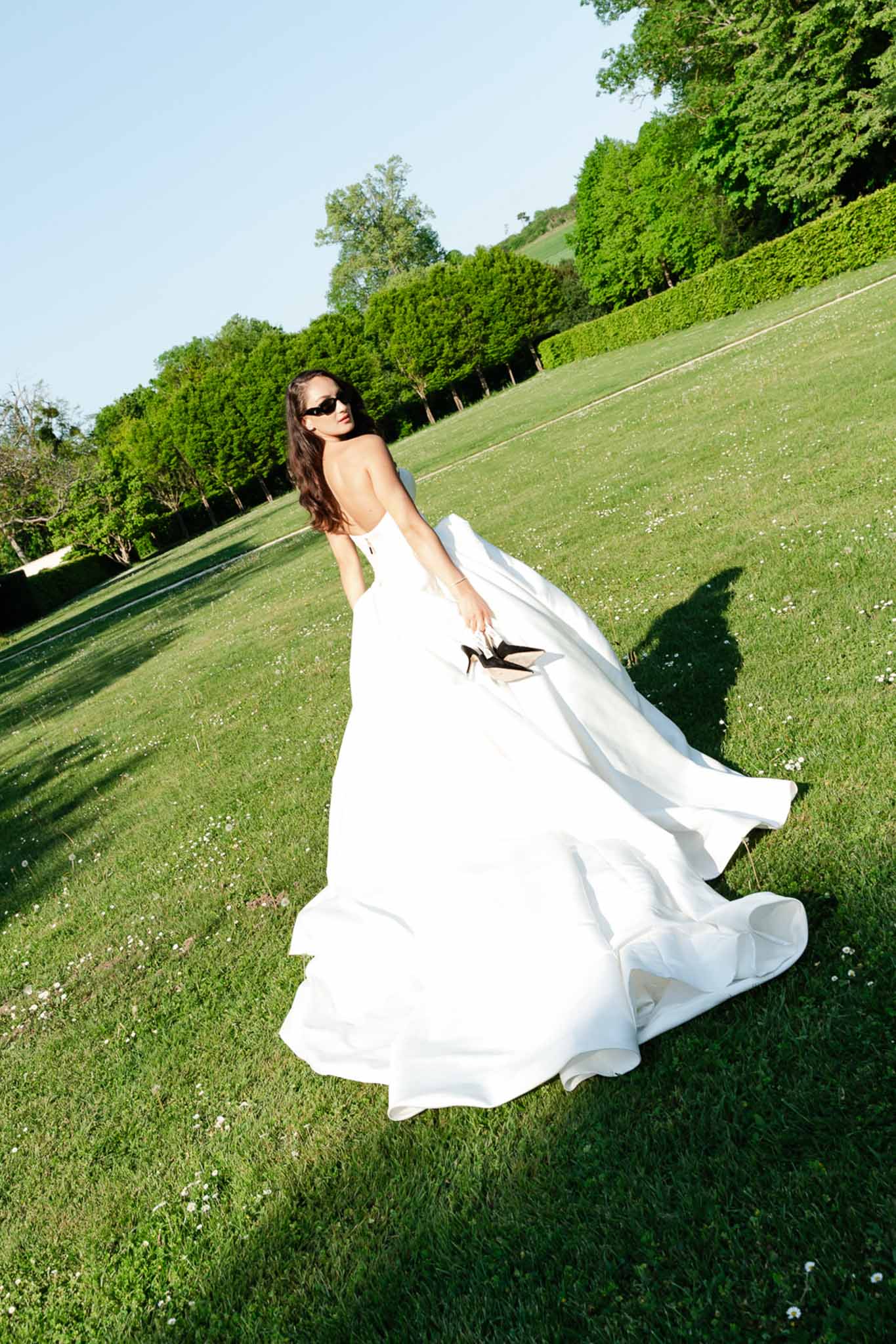 Bride walking barefoot on lawn in strapless ball gown carrying black heels and wearing cat-eye sunglasses