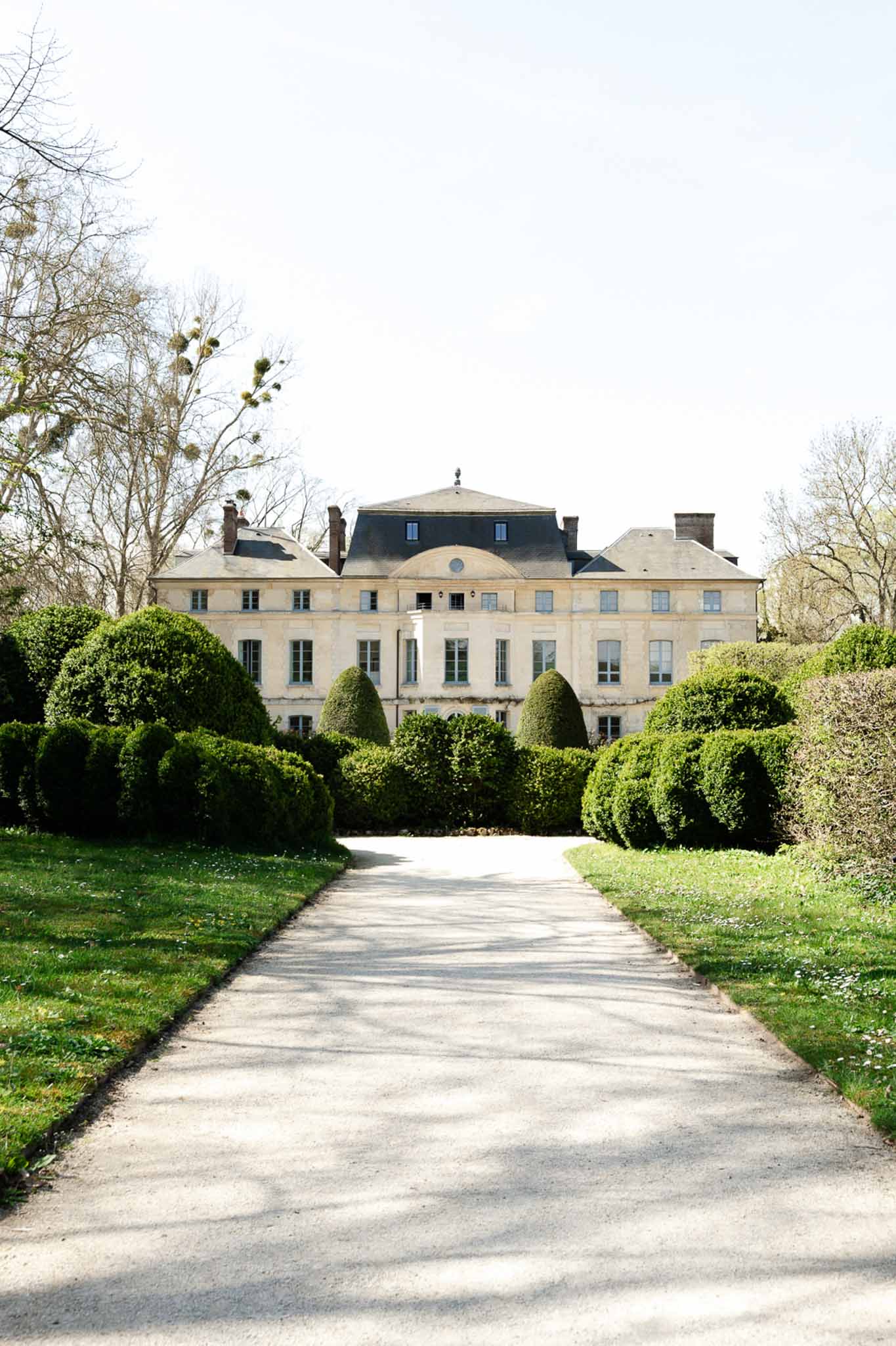 Symmetrical gravel driveway flanked by trimmed topiary leading to cream stone chateau