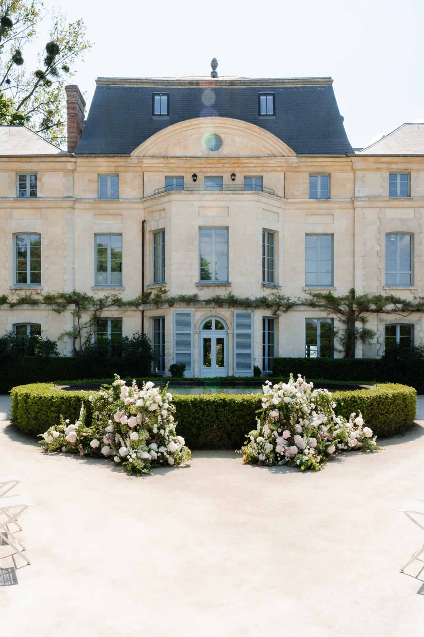 Two large blush and white floral arrangements at curved boxwood hedge before limestone chateau facade