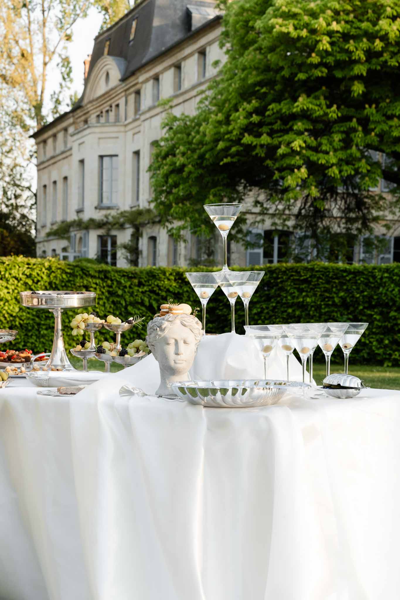 Cocktail hour display table with martini glass tower, classical bust, and silver tiered stands at French chateau