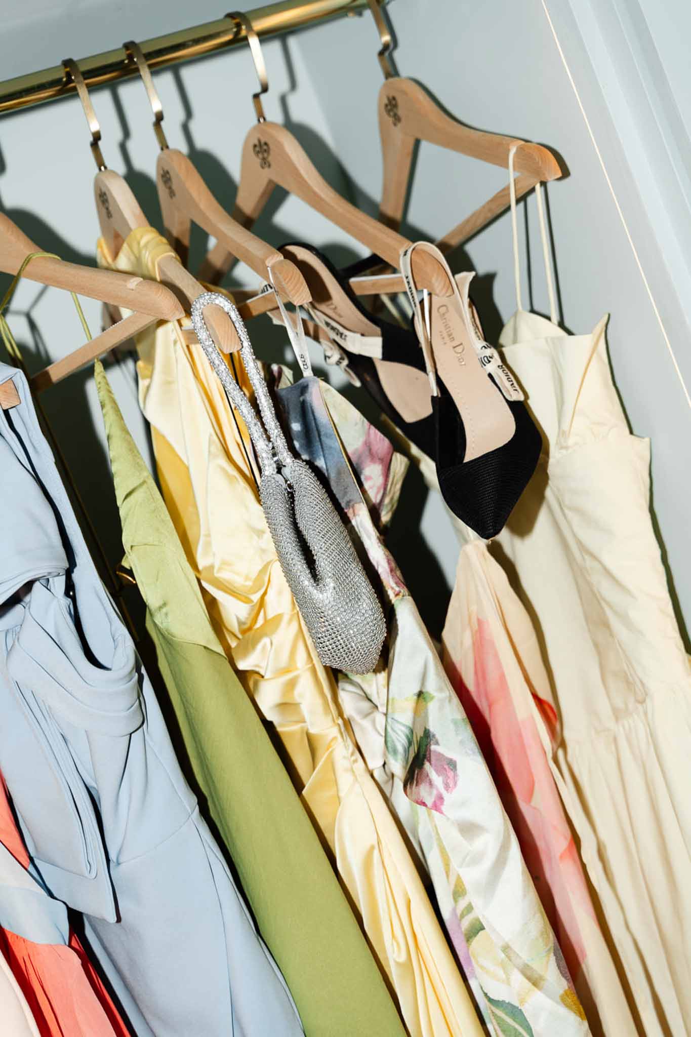 Wedding outfits on brass rail with blue, sage, and yellow dresses, Dior heels, and crystal evening bag