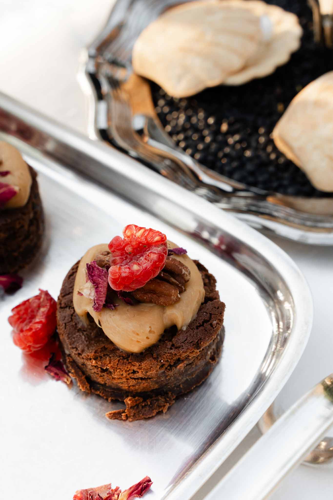 Chocolate brownie bite with mousse, pecan, raspberry, and dried rose petals on silver tray beside caviar