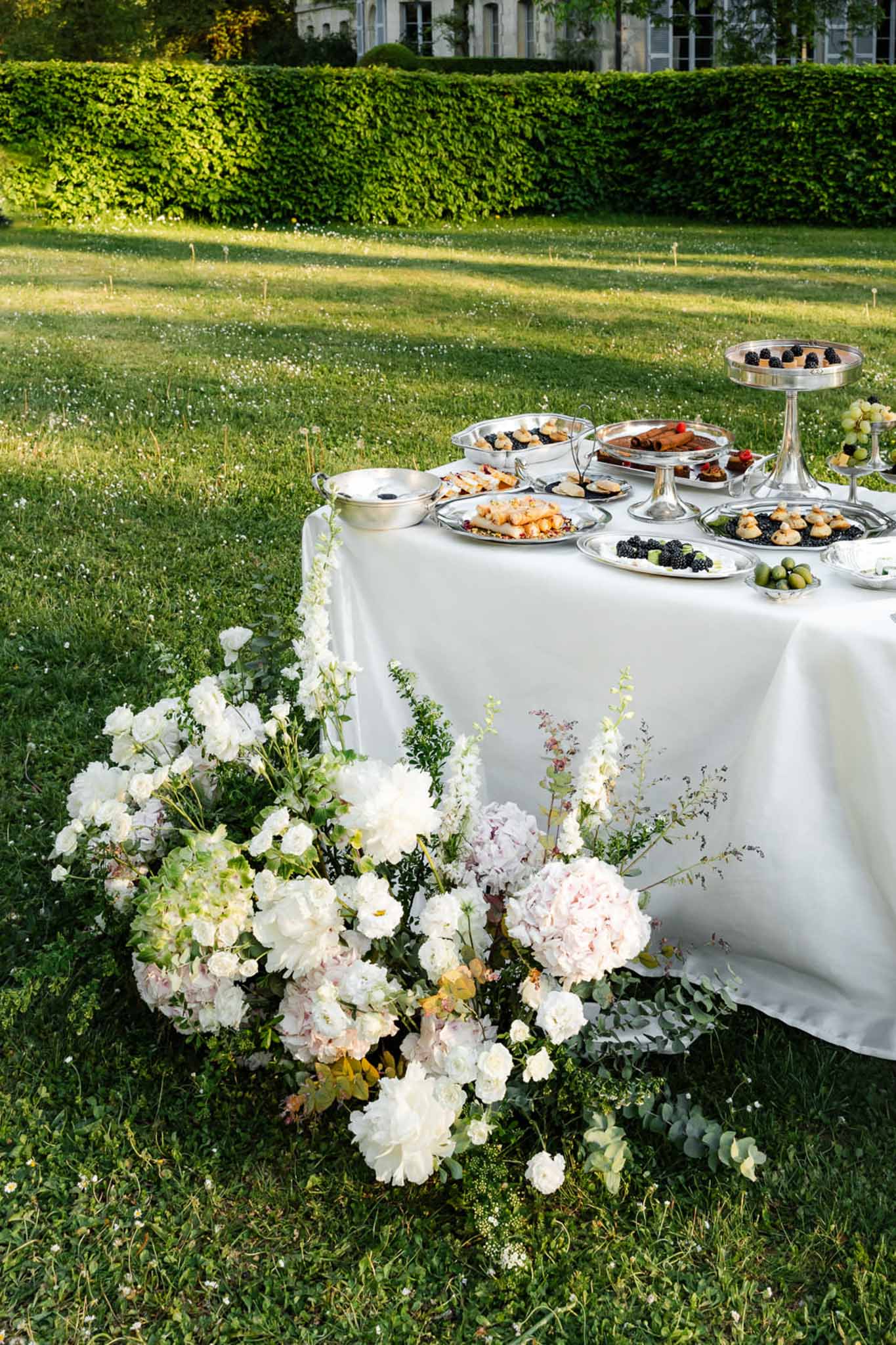 Silver canape platters on white linen table with white rose and hydrangea ground arrangement on lawn
