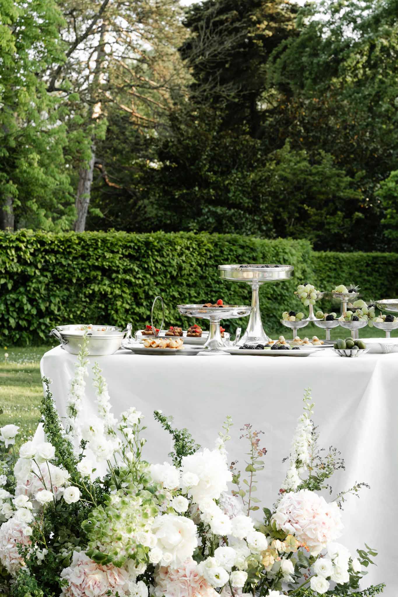 Silver tiered canape display with white ranunculus and blush peony ground arrangement in formal garden