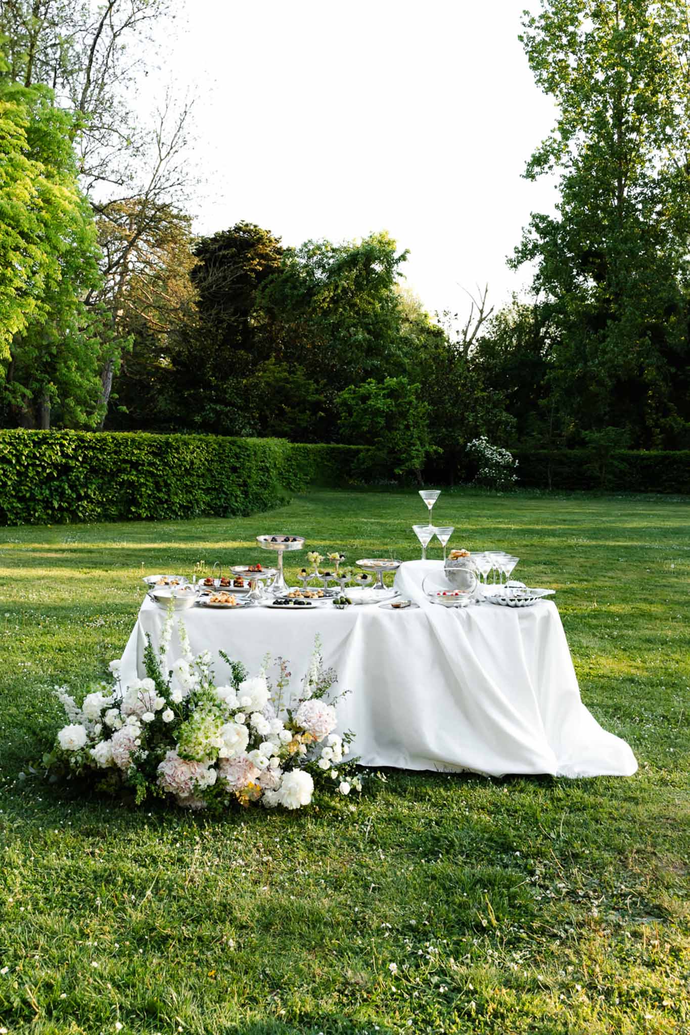 Dessert table with silver tiered stands and white peony ground arrangement in formal garden
