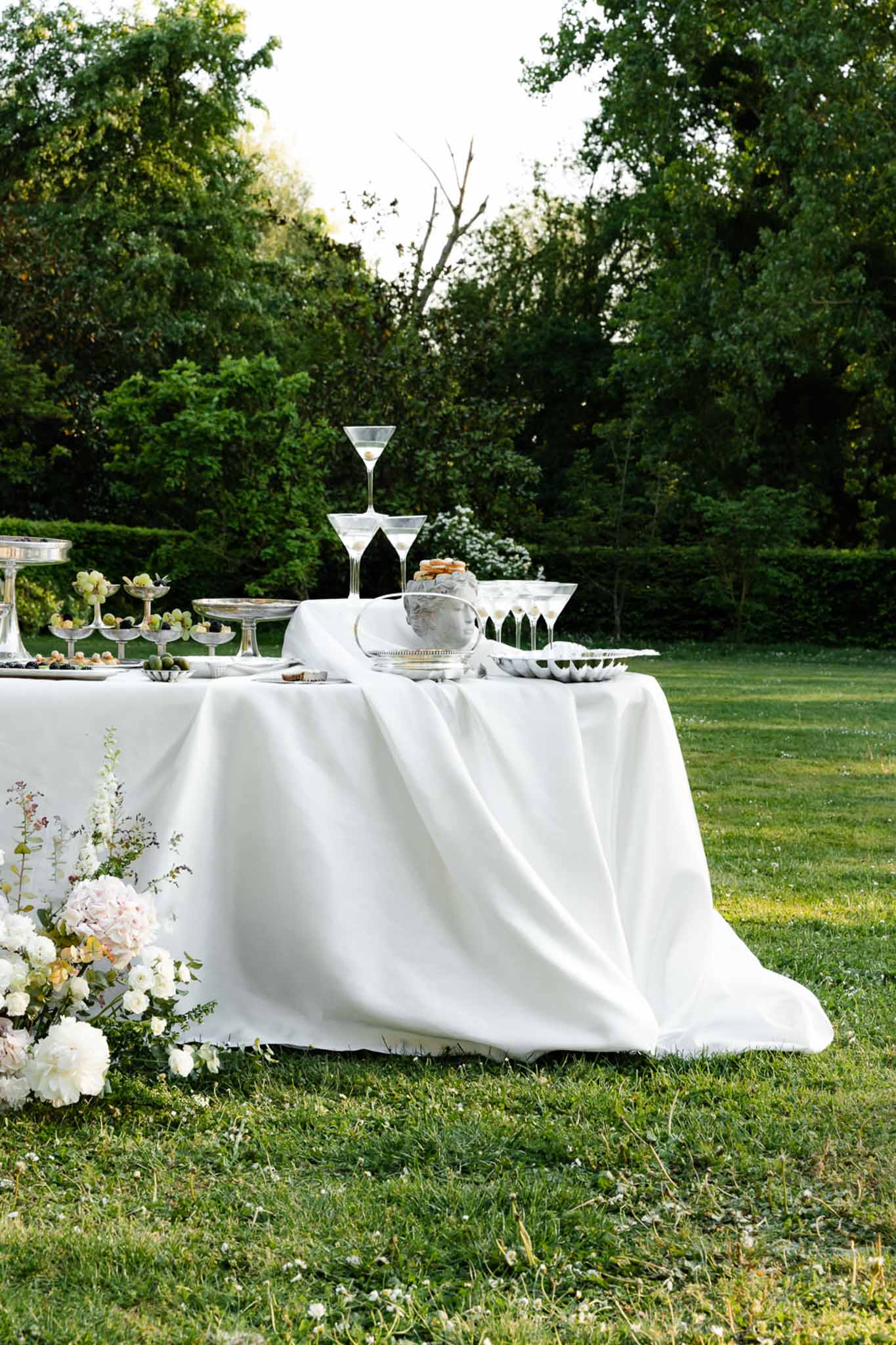 Outdoor cocktail table with silver stands, fresh fruit, coupe glasses, and white peony floral arrangement