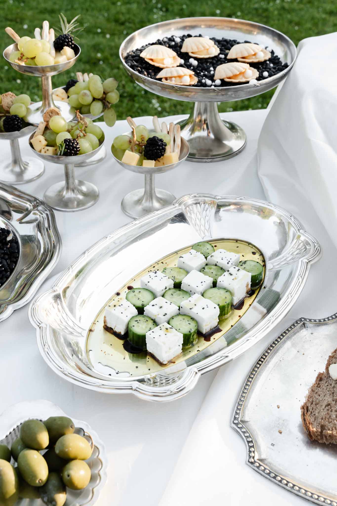 Silver platters with cheese, olives, grapes, bread, and confections on white linen at outdoor cocktail display