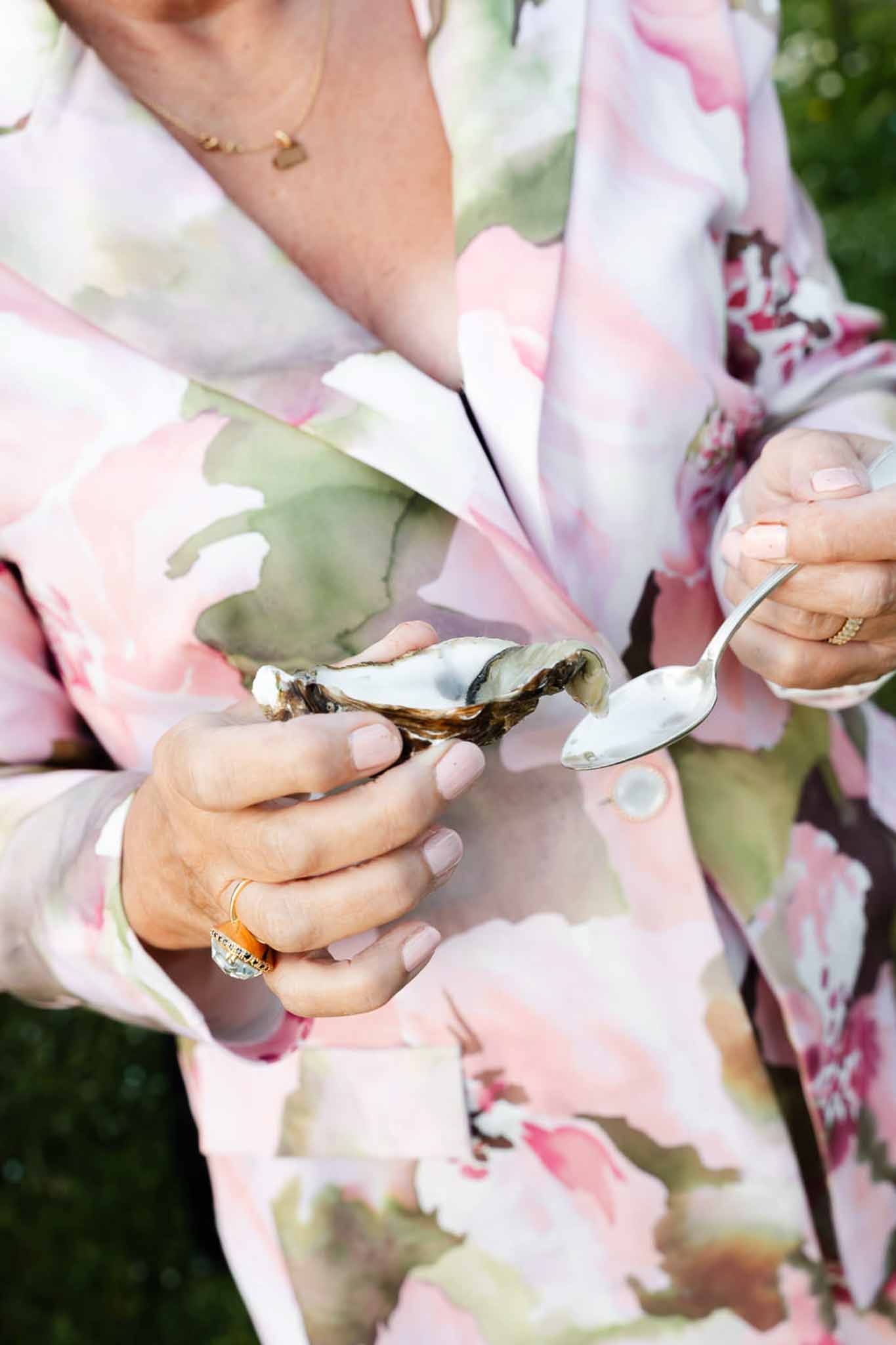 Close-up of guest eating oyster in floral-print blazer with gold rings at outdoor garden cocktail hour