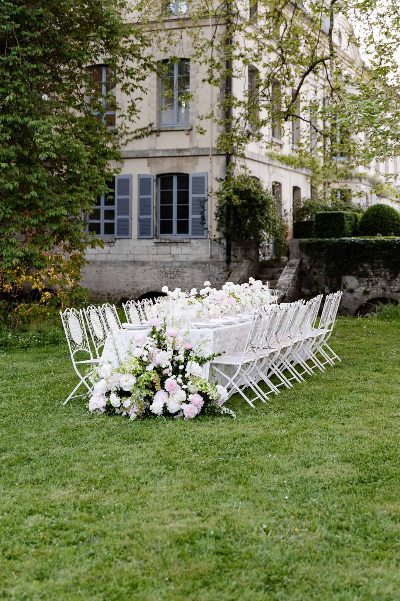 Long outdoor reception table with blush peony centerpieces and white wrought-iron chairs on chateau lawn