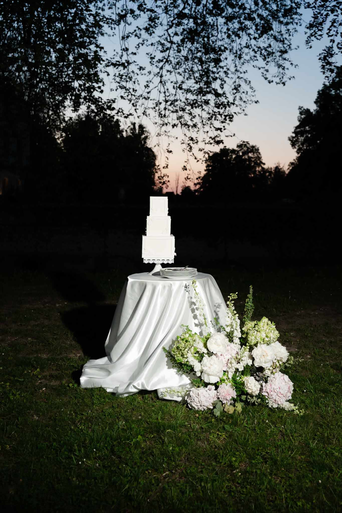 Four-tier white cake on silver stand with cascading blush hydrangea and peony arrangement at dusk