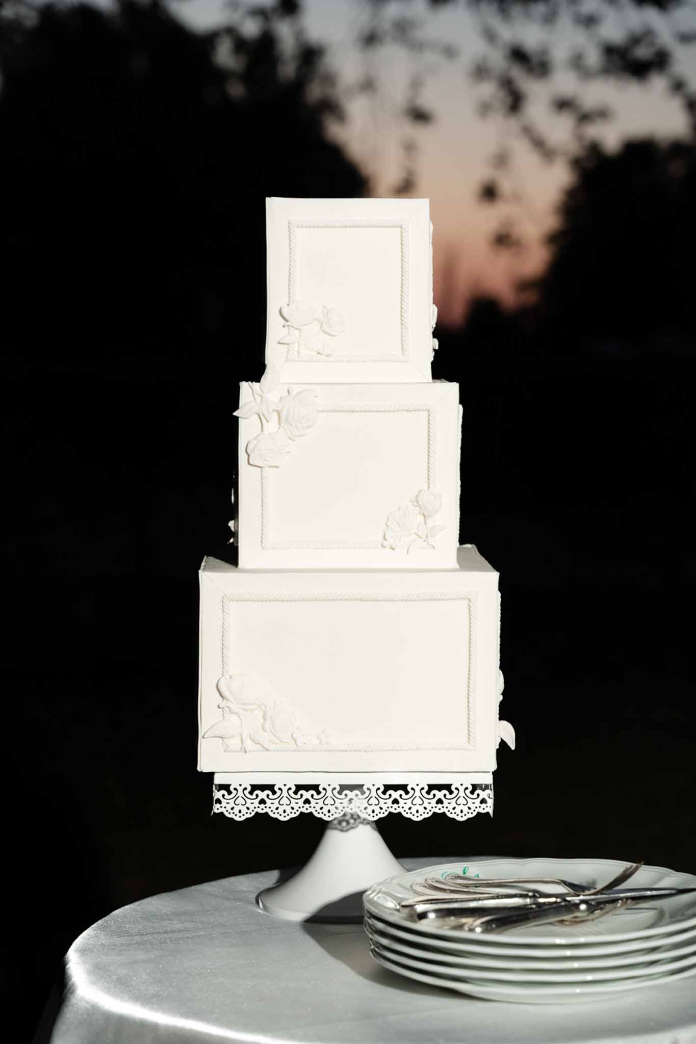 Three-tier square white fondant wedding cake with sugar floral appliques on a laser-cut stand at dusk