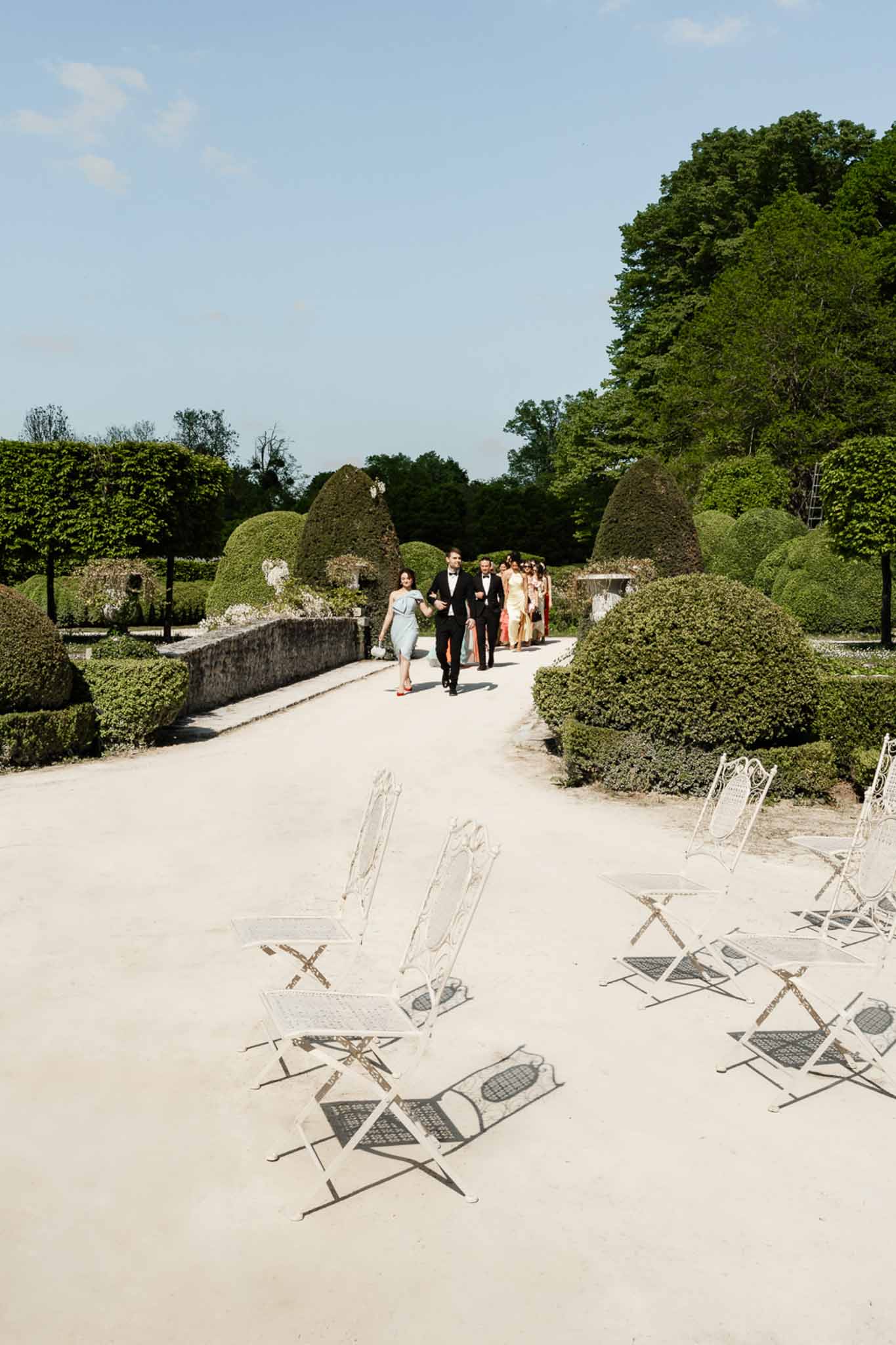 Wedding guests and bridal party walking along gravel path through formal French chateau garden toward ceremony chairs