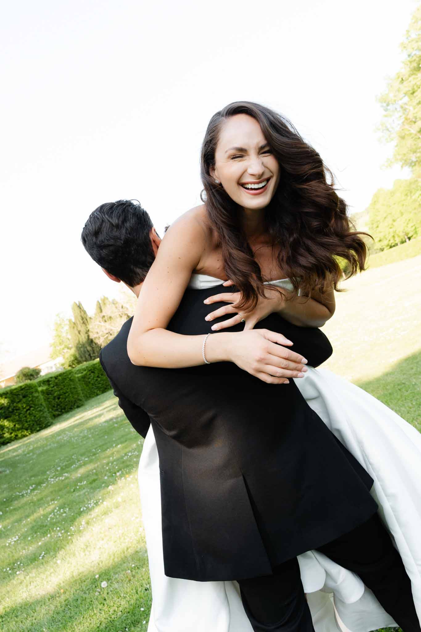Groom lifting laughing bride in strapless white gown in a manicured garden setting