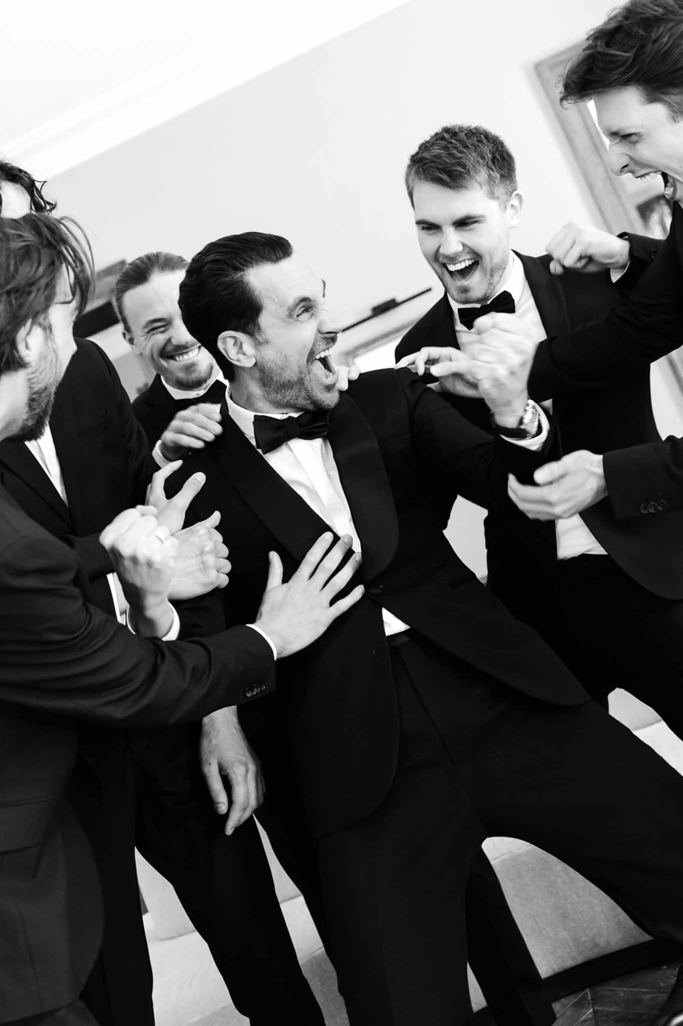 Groom and four groomsmen in tuxedos laughing and roughhousing in high-energy indoor portrait in B&W