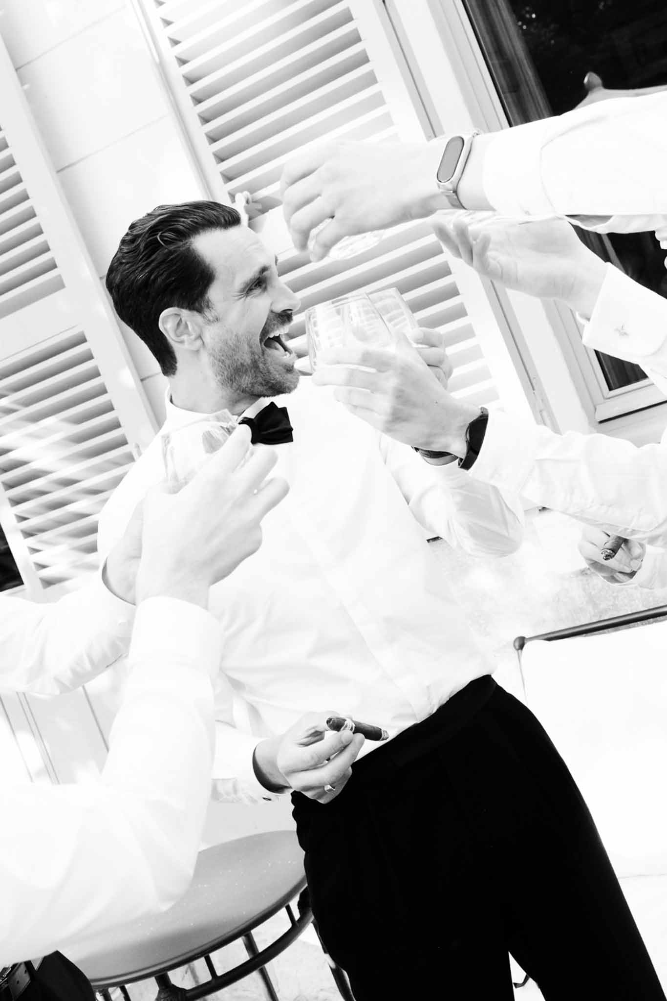 Groom laughing with cigar and reaching for glass in white shirt and bow tie during reception in B&W