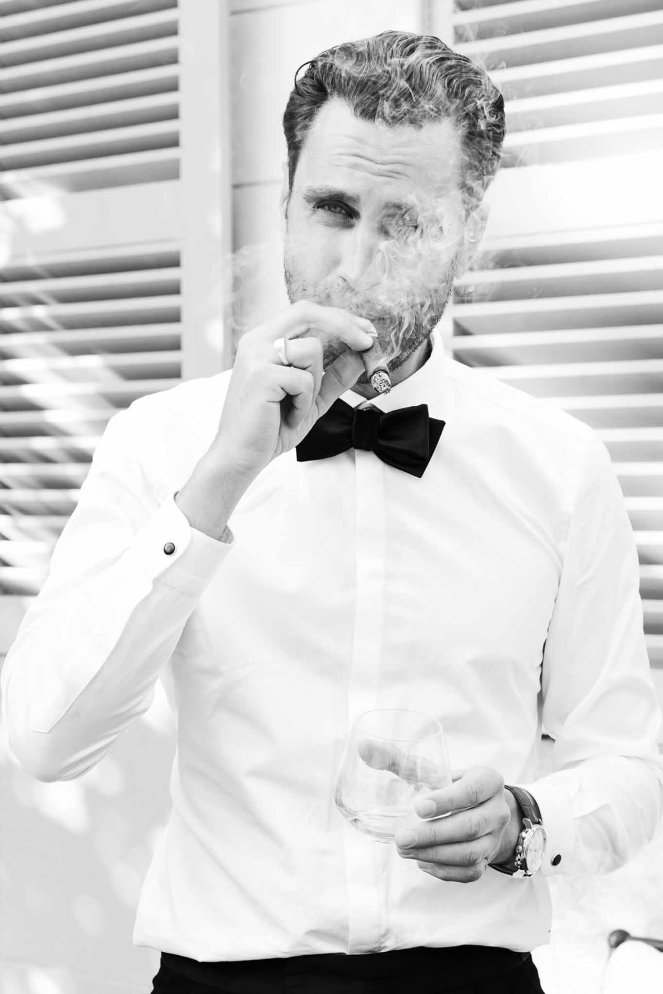 Black-and-white portrait of groom in bow tie and French cuffs smoking a cigar and holding a glass