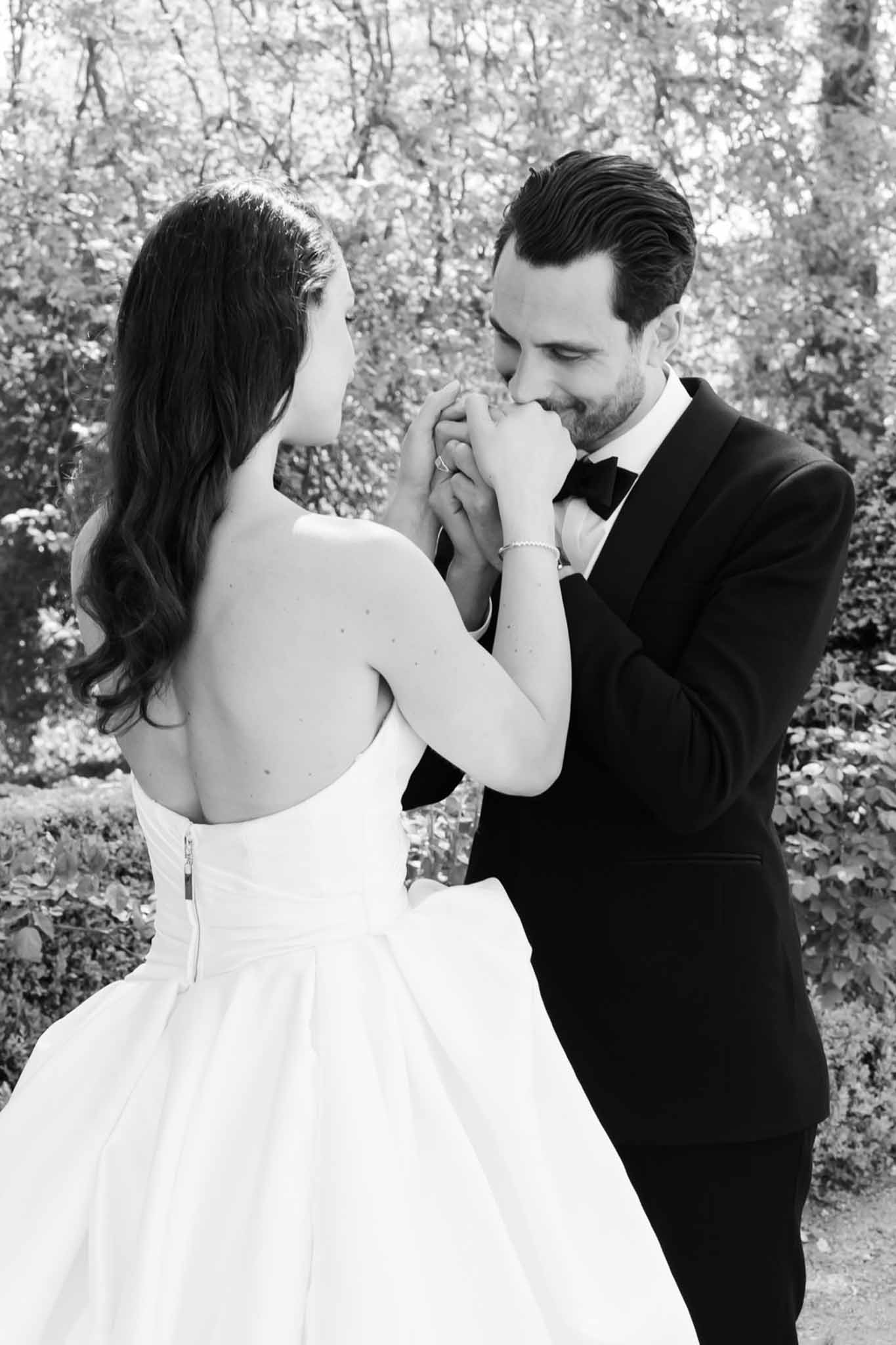Black and white portrait of groom kissing bride's clasped hands in garden setting