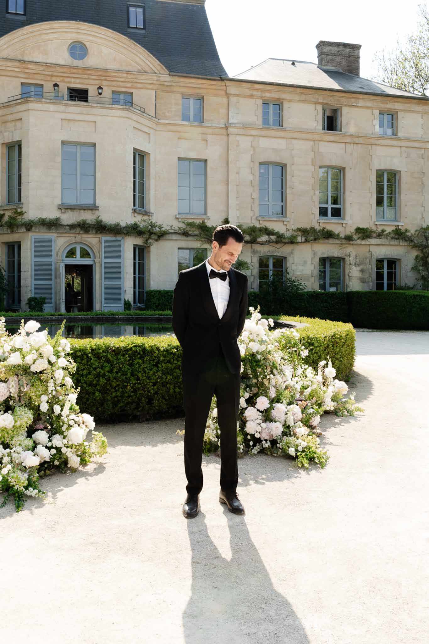 Groom in black tuxedo between white peony ceremony arrangements before limestone chateau with reflecting pool