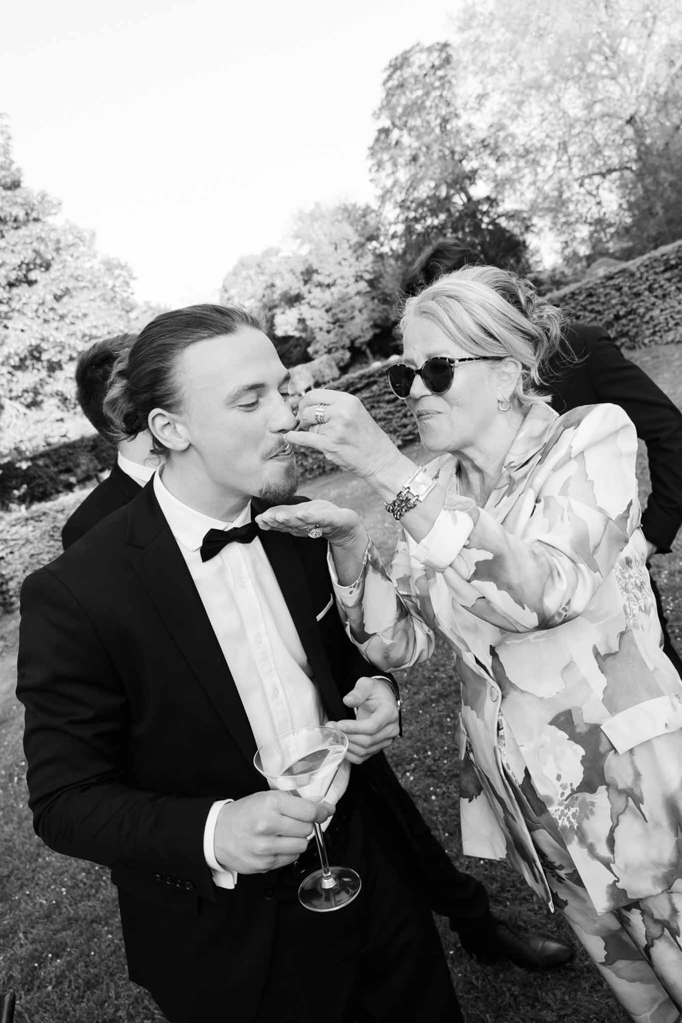 Man in tuxedo kissing hand of woman in floral dress during garden cocktail hour in B&W