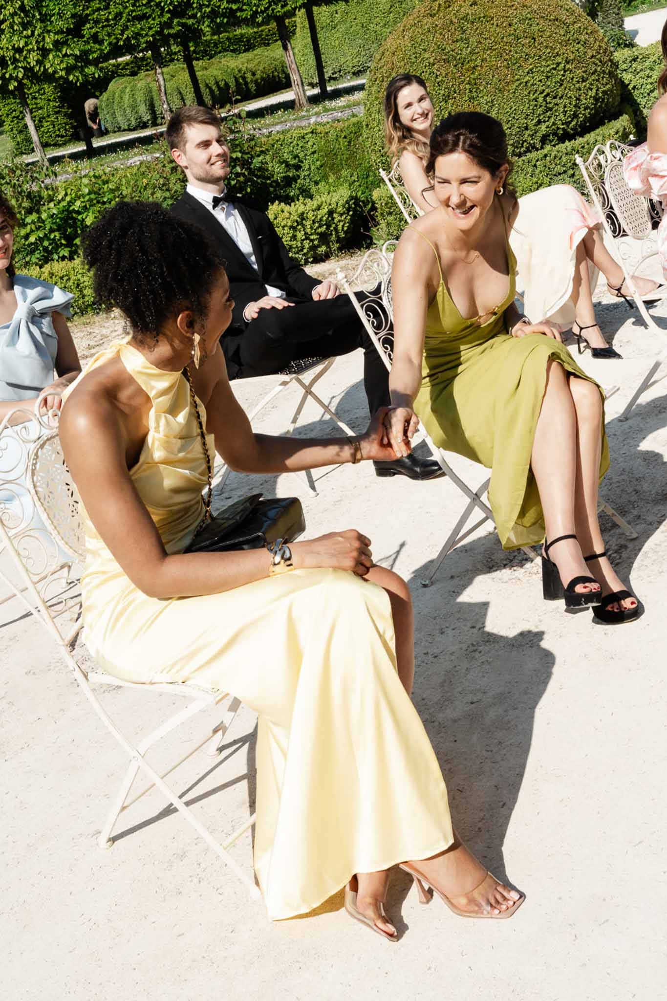Wedding guests laughing on white wrought-iron chairs in formal chateau garden during cocktail hour