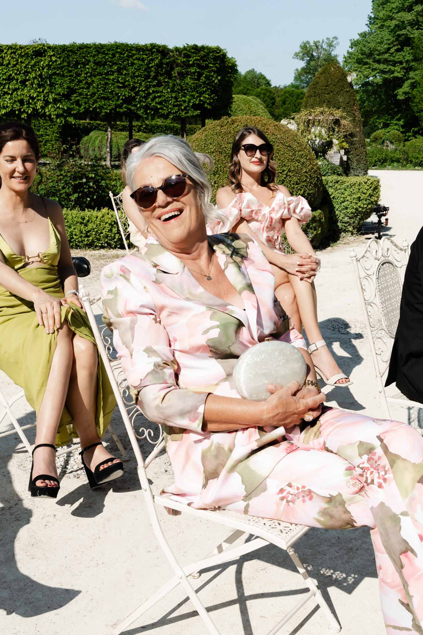 Three female guests in floral pink and chartreuse outfits laughing on wrought iron garden chairs in sunlight
