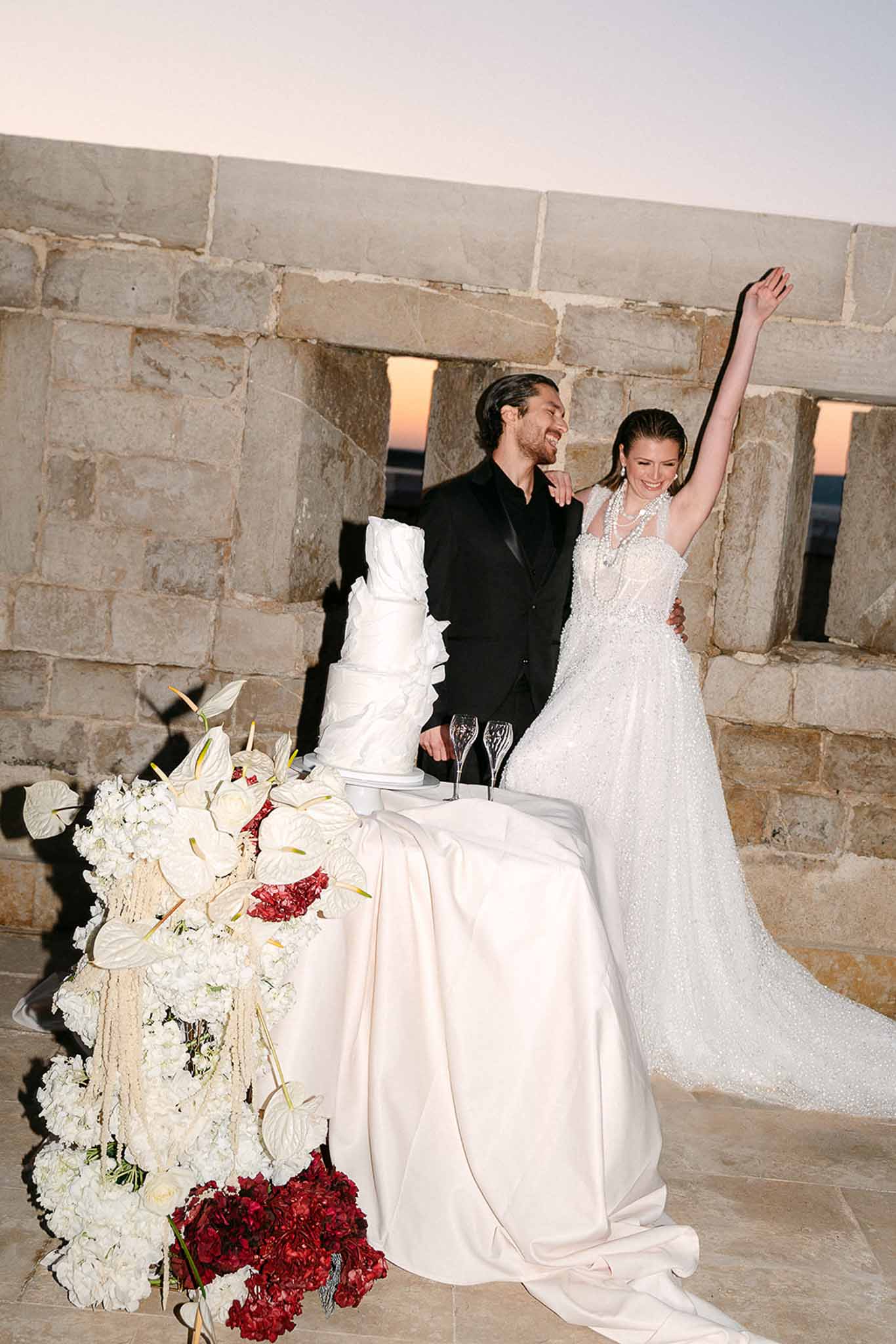 Couple celebrating after cake cutting with four-tier white cake and floral arrangement at dusk terrace