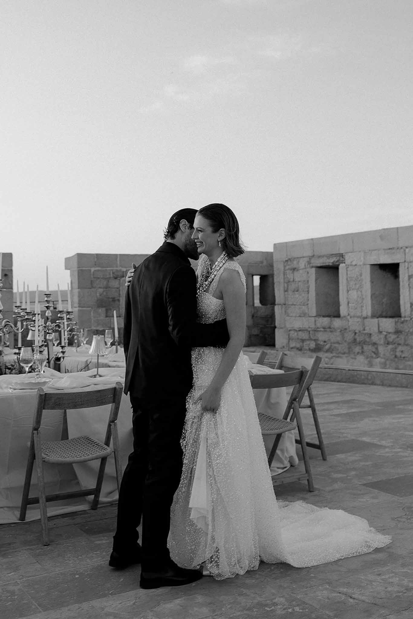 Bride in beaded gown laughing as groom leans in on a rooftop terrace reception setting in black and white