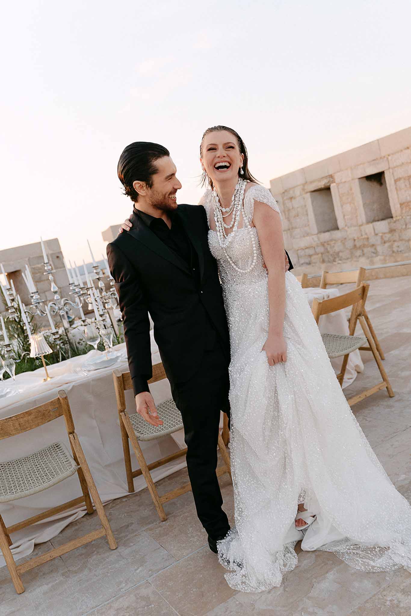 Bride in beaded ivory gown and groom in all-black suit laughing on stone terrace near reception table