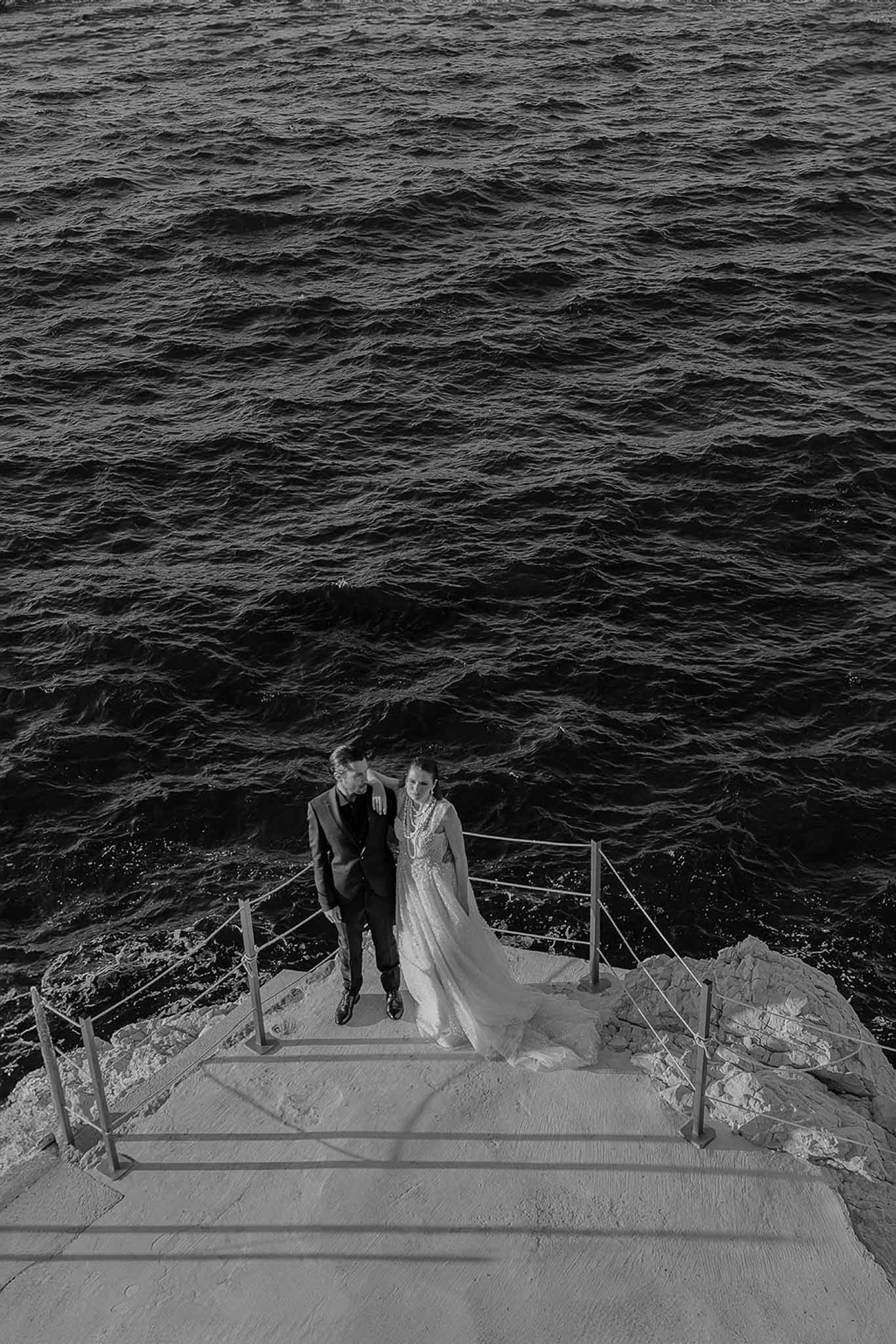 Aerial black-and-white portrait of bride and groom standing on a stone jetty over dark rippling water