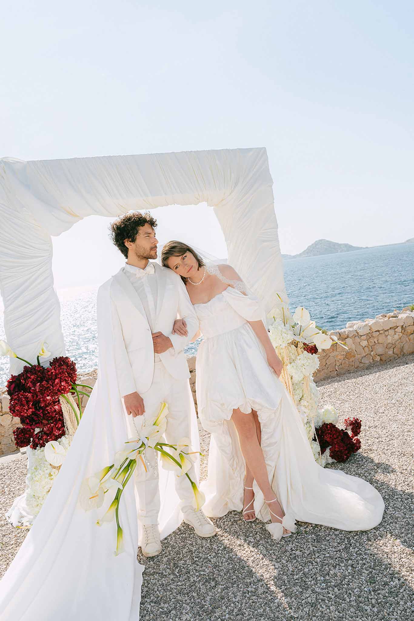 Couple posing before draped ivory ceremony arch with burgundy hydrangeas and white orchids on coastal terrace