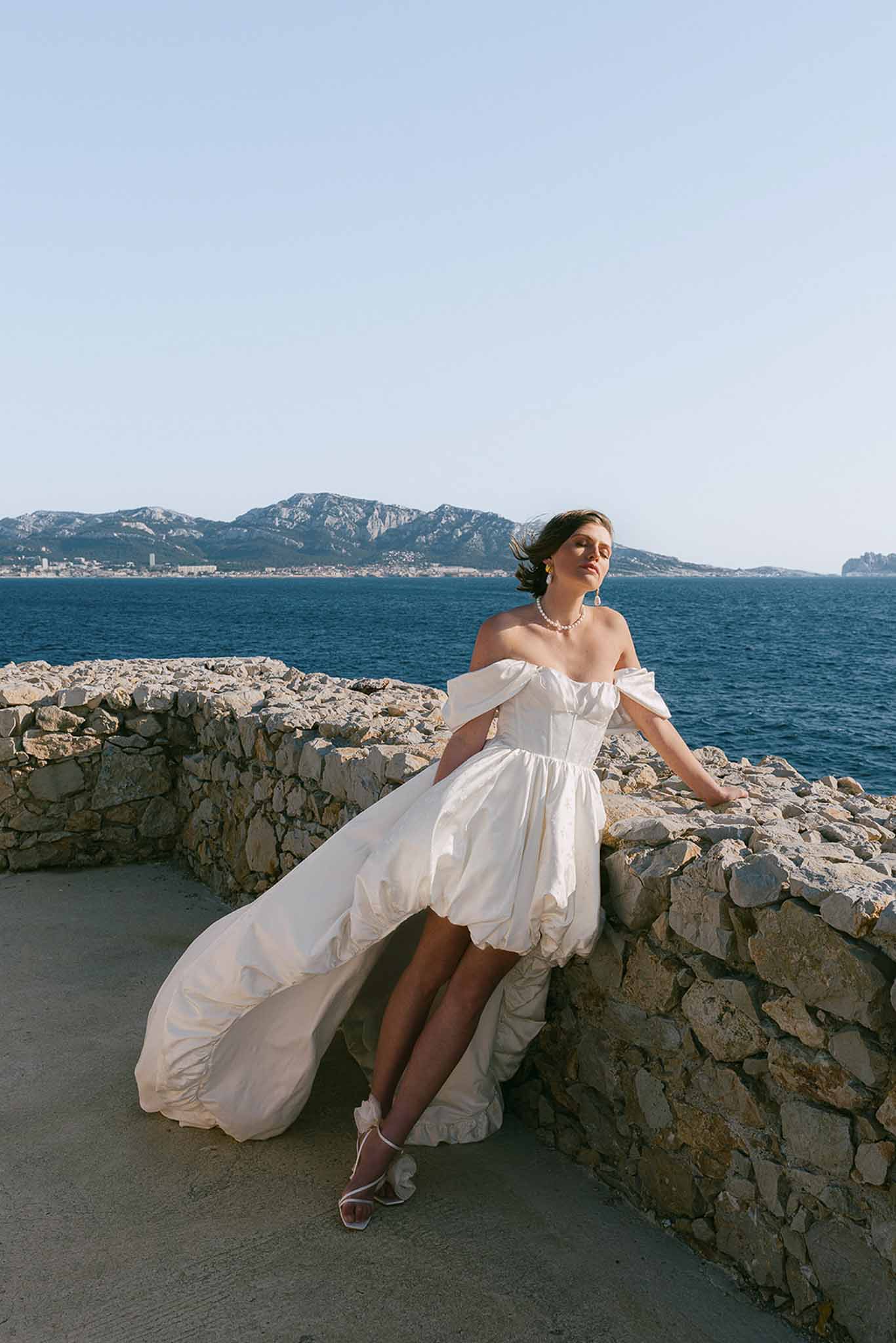 Bride in off-white taffeta bubble hem gown leaning on sea wall with Mediterranean coastline and mountains behind