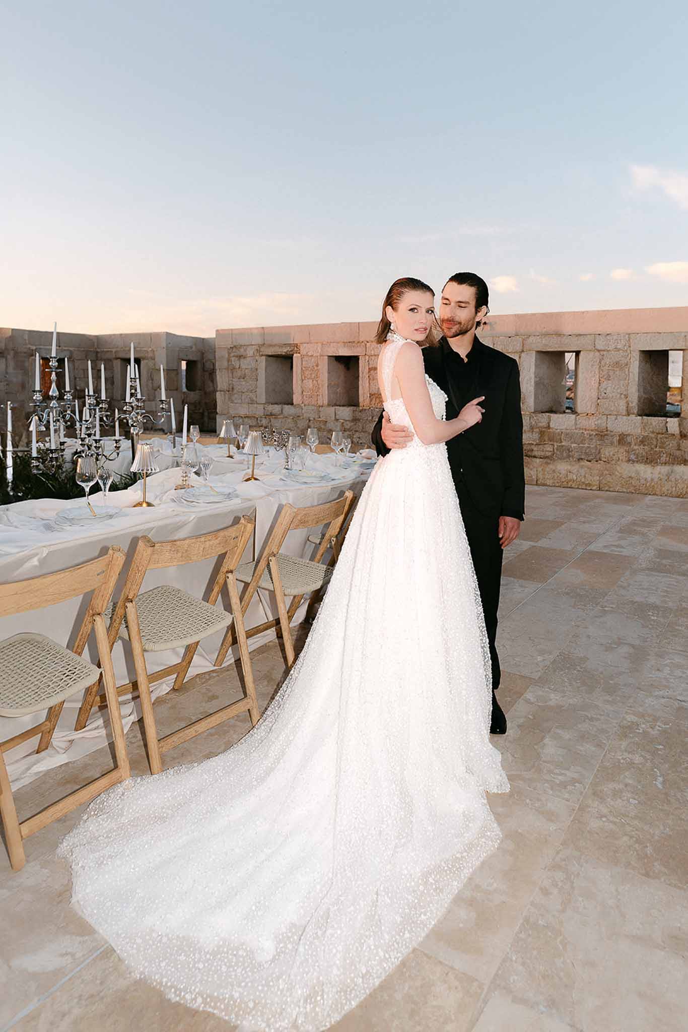 Bride with embellished train and groom on castle rooftop terrace beside blue and white table setting