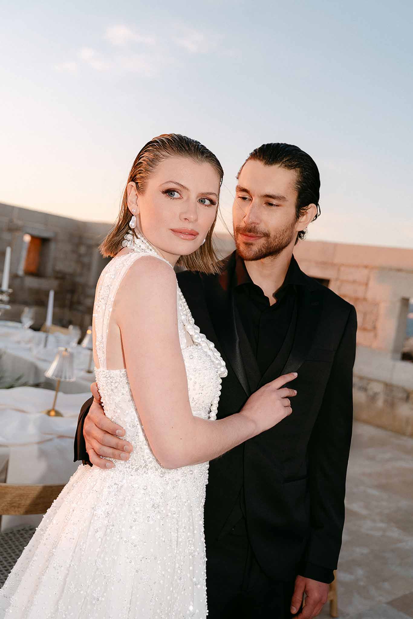 Couple at dusk on stone terrace, bride in beaded pearl gown with slicked hair, groom in all-black tuxedo