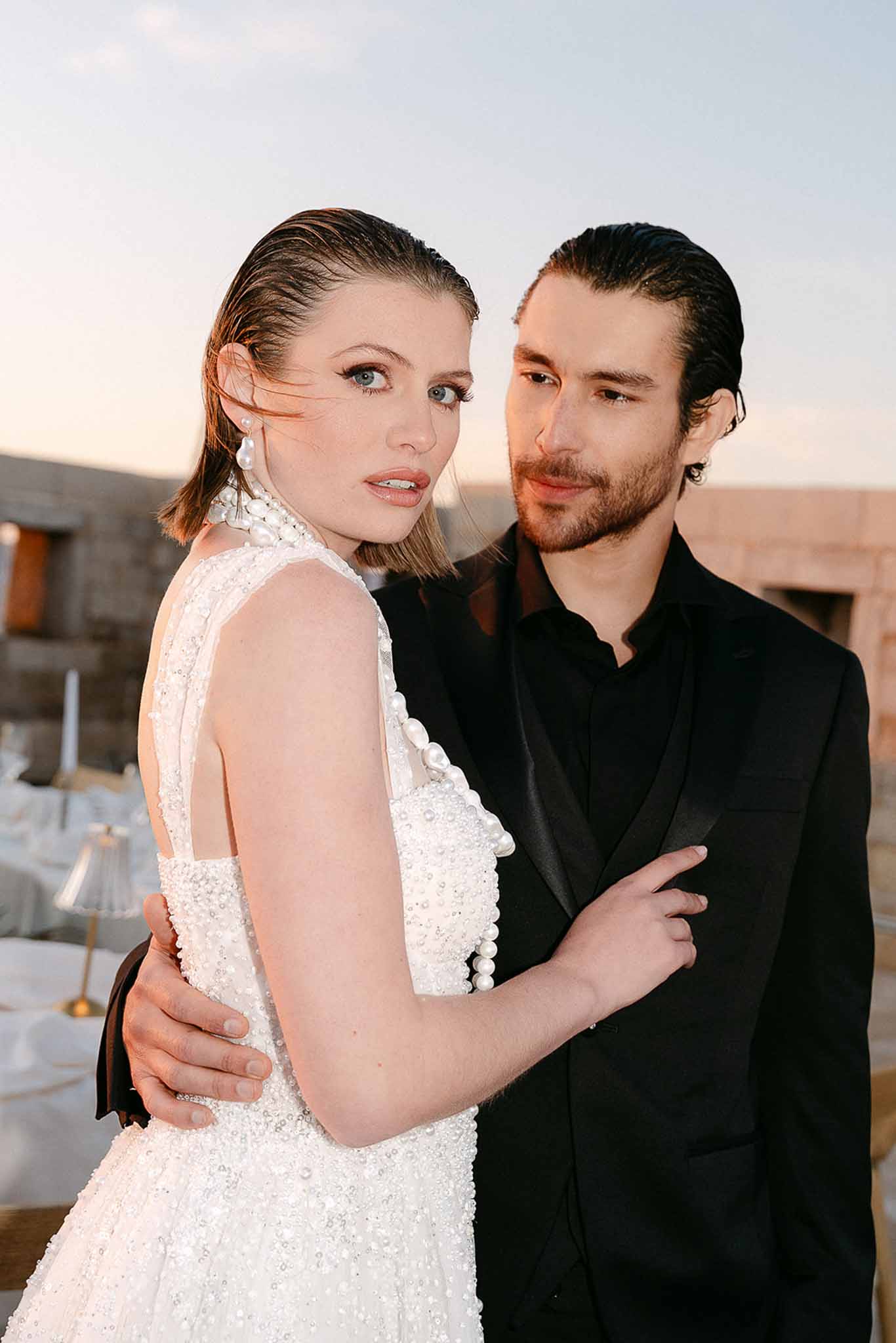 Close-up of bride in crystal-beaded gown with pearl choker beside groom in all-black at golden hour