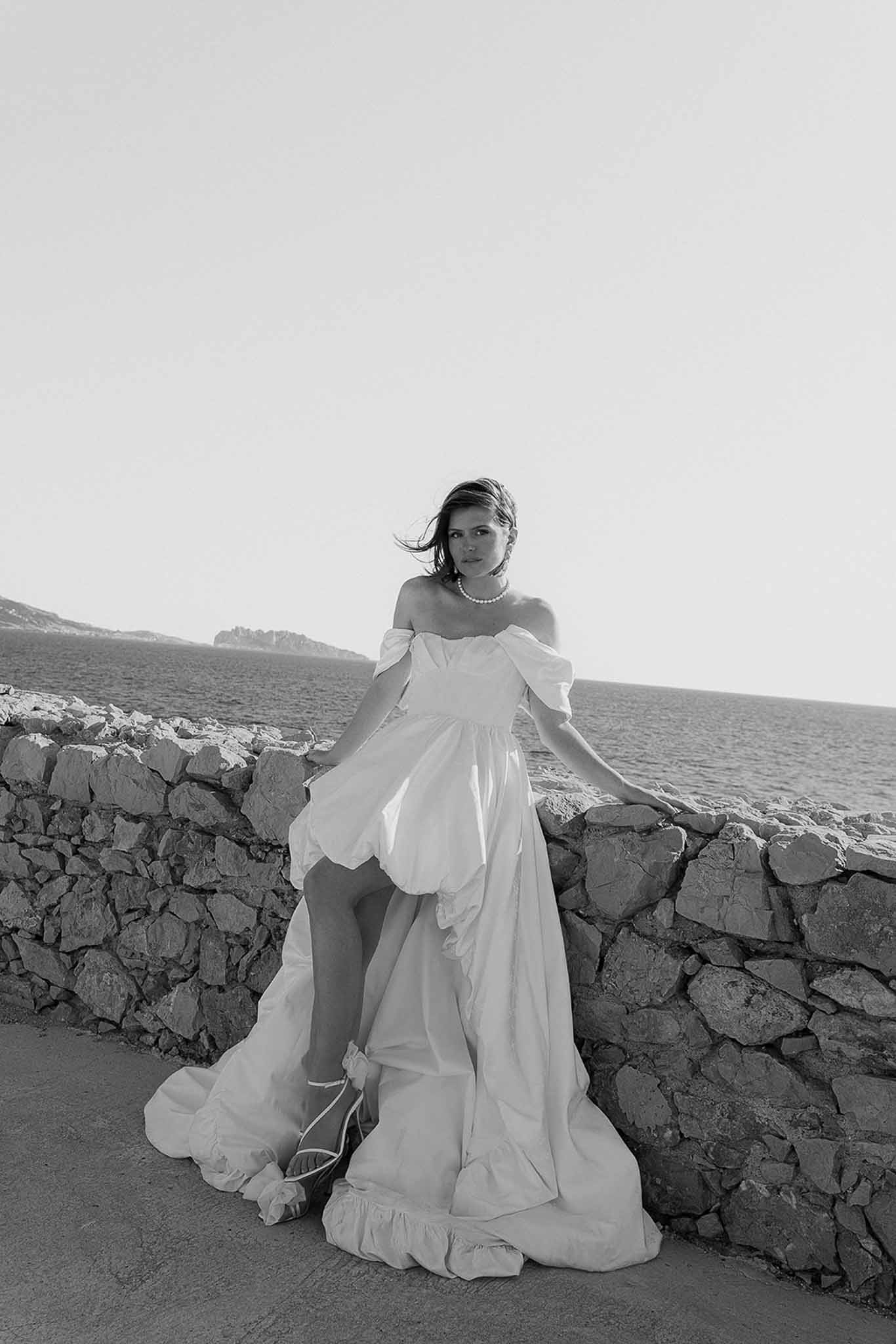 Black-and-white portrait of bride in off-the-shoulder gown leaning against stone wall with sea view