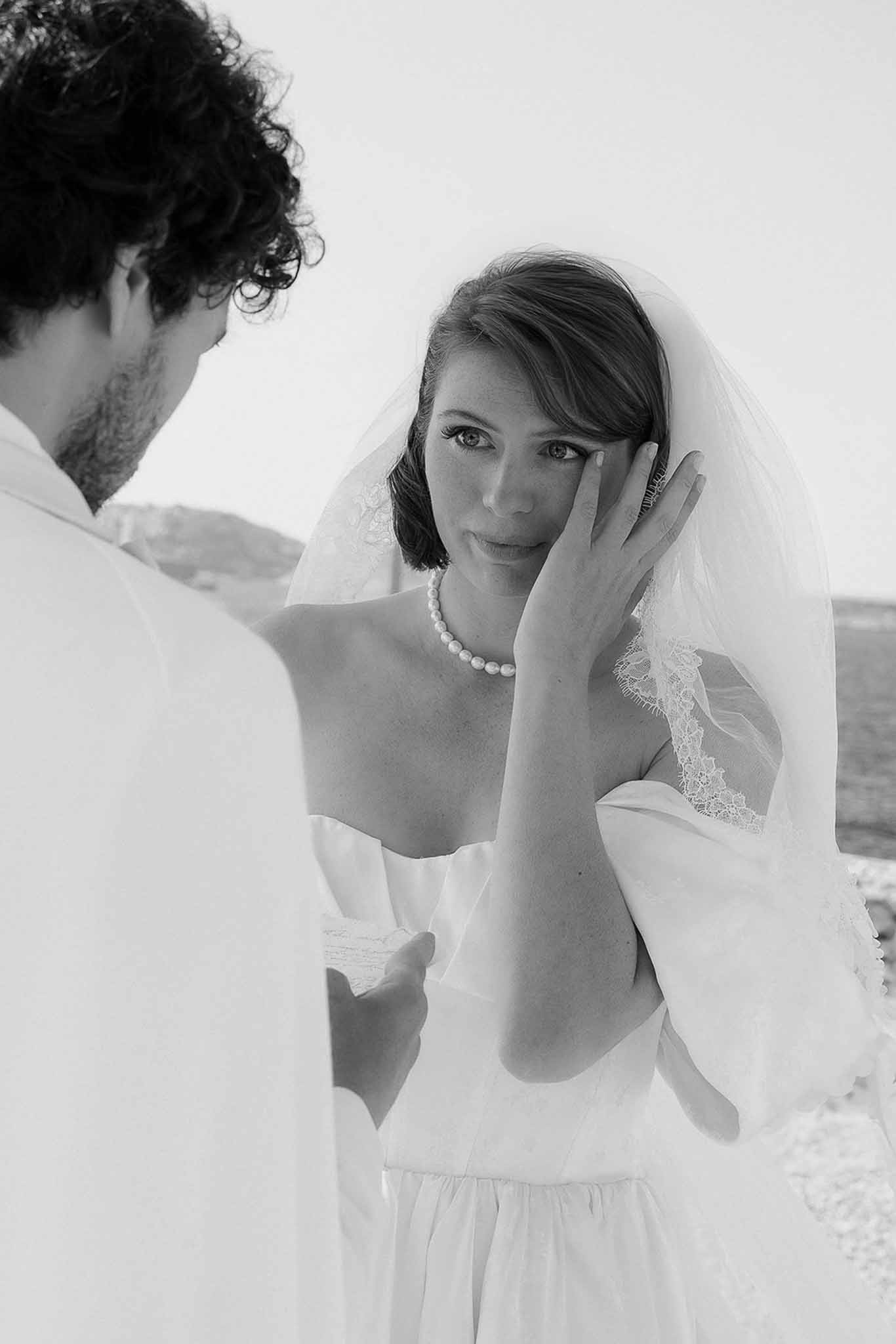 Black and white close-up of smiling bride with veil and pearl necklace framed over groom shoulder