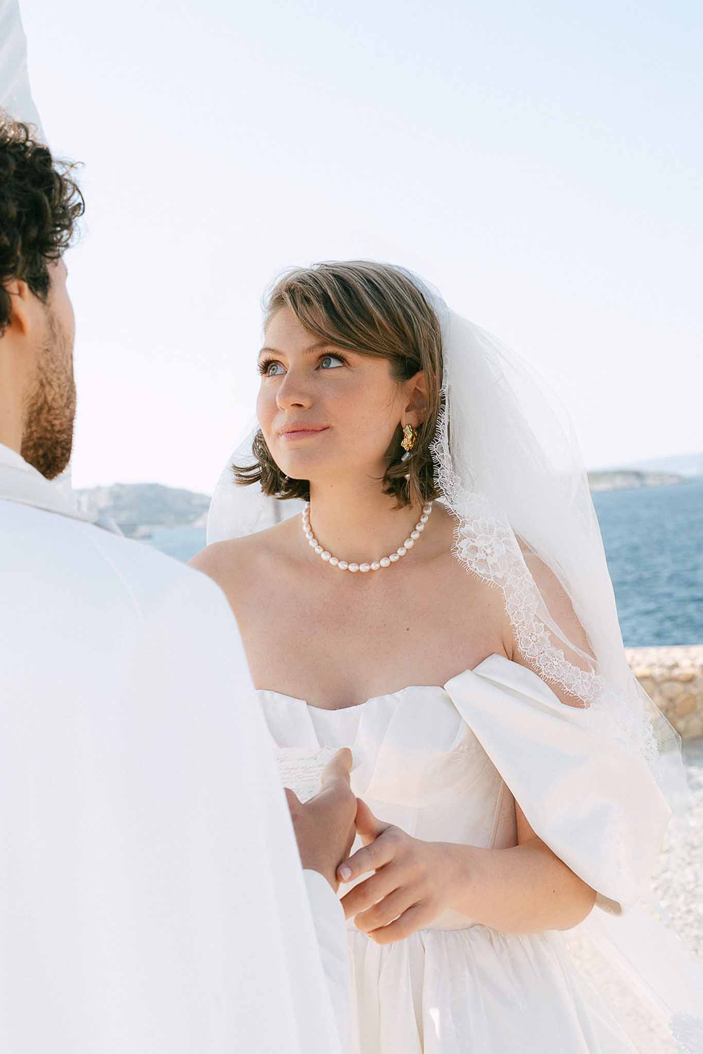 Close-up of bride in strapless gown with puff sleeves, pearl choker, and gold earrings facing groom at coastal location