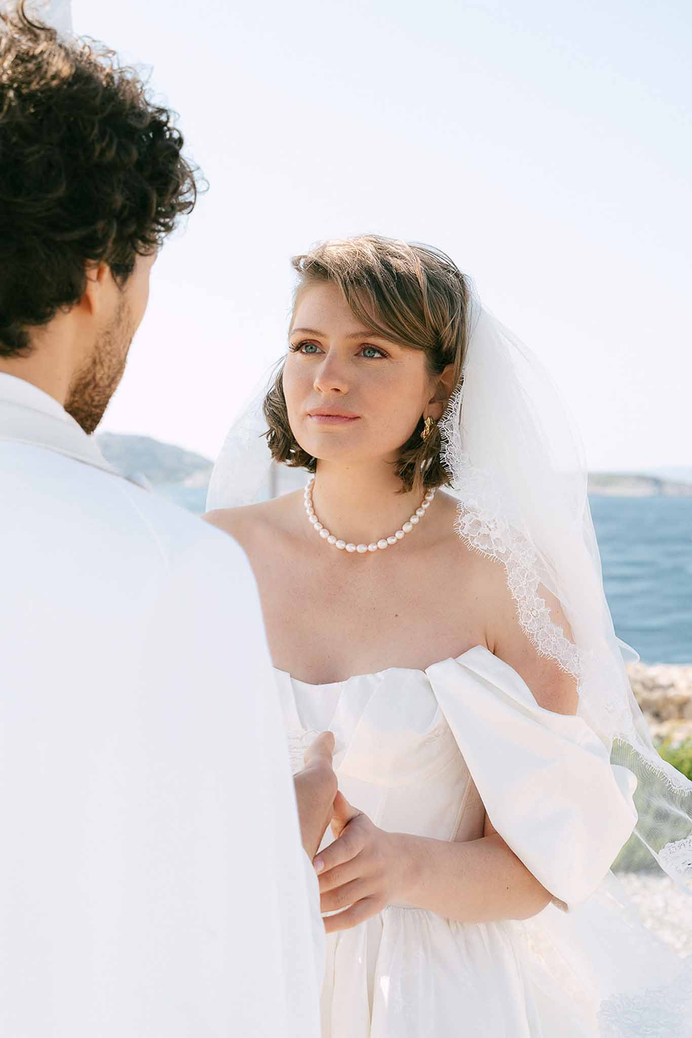 Close-up of bride in pearl choker and lace veil looking at groom during coastal ceremony