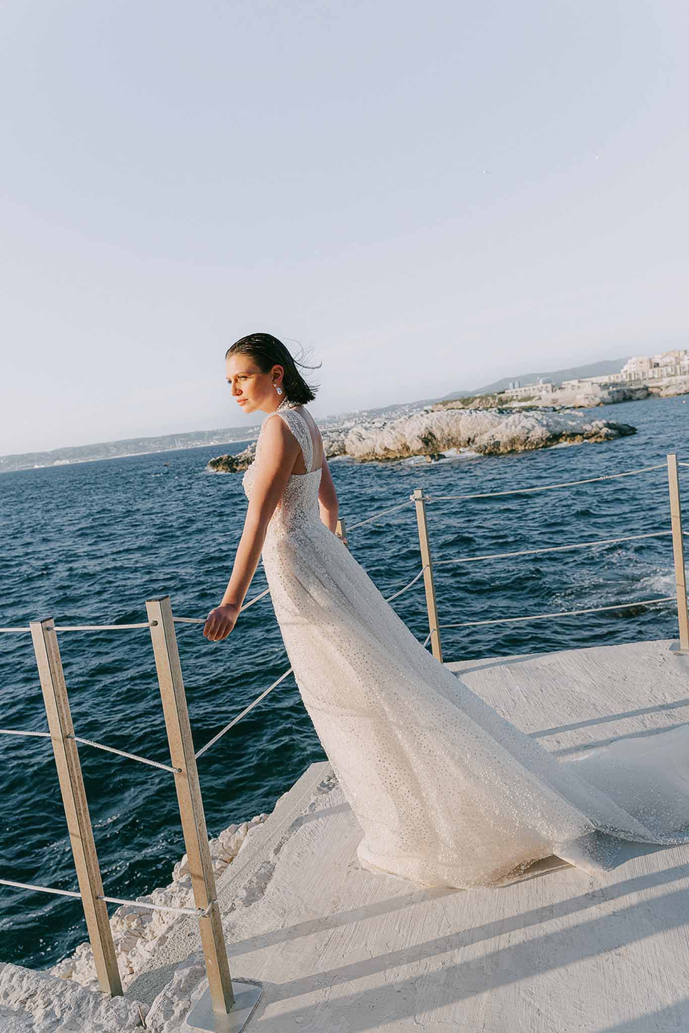 Bride in beaded ivory gown looks over shoulder on white seaside terrace with Mediterranean coast beyond