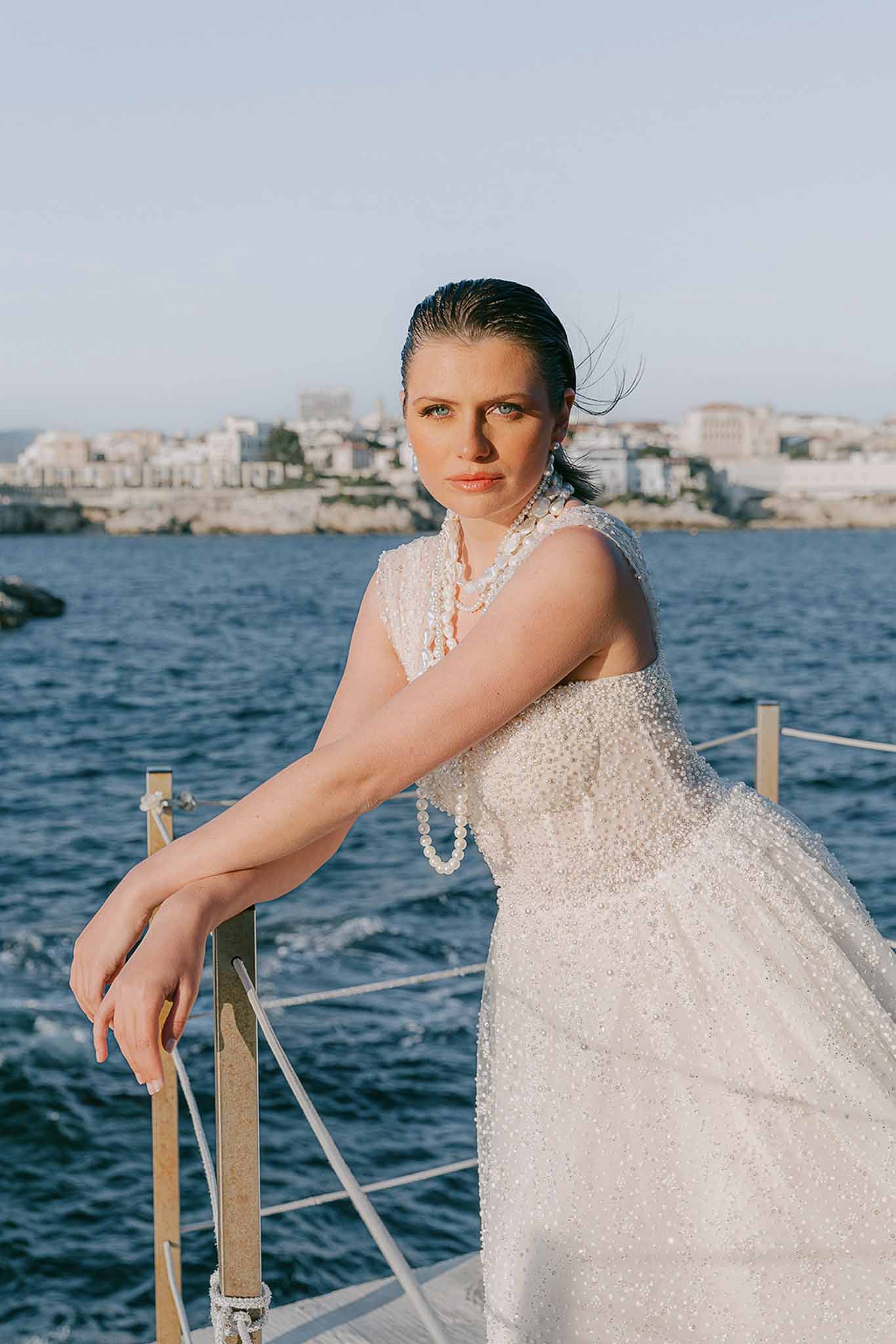 Bride in beaded ivory gown with layered pearl necklaces leaning on boat railing with coastal town backdrop