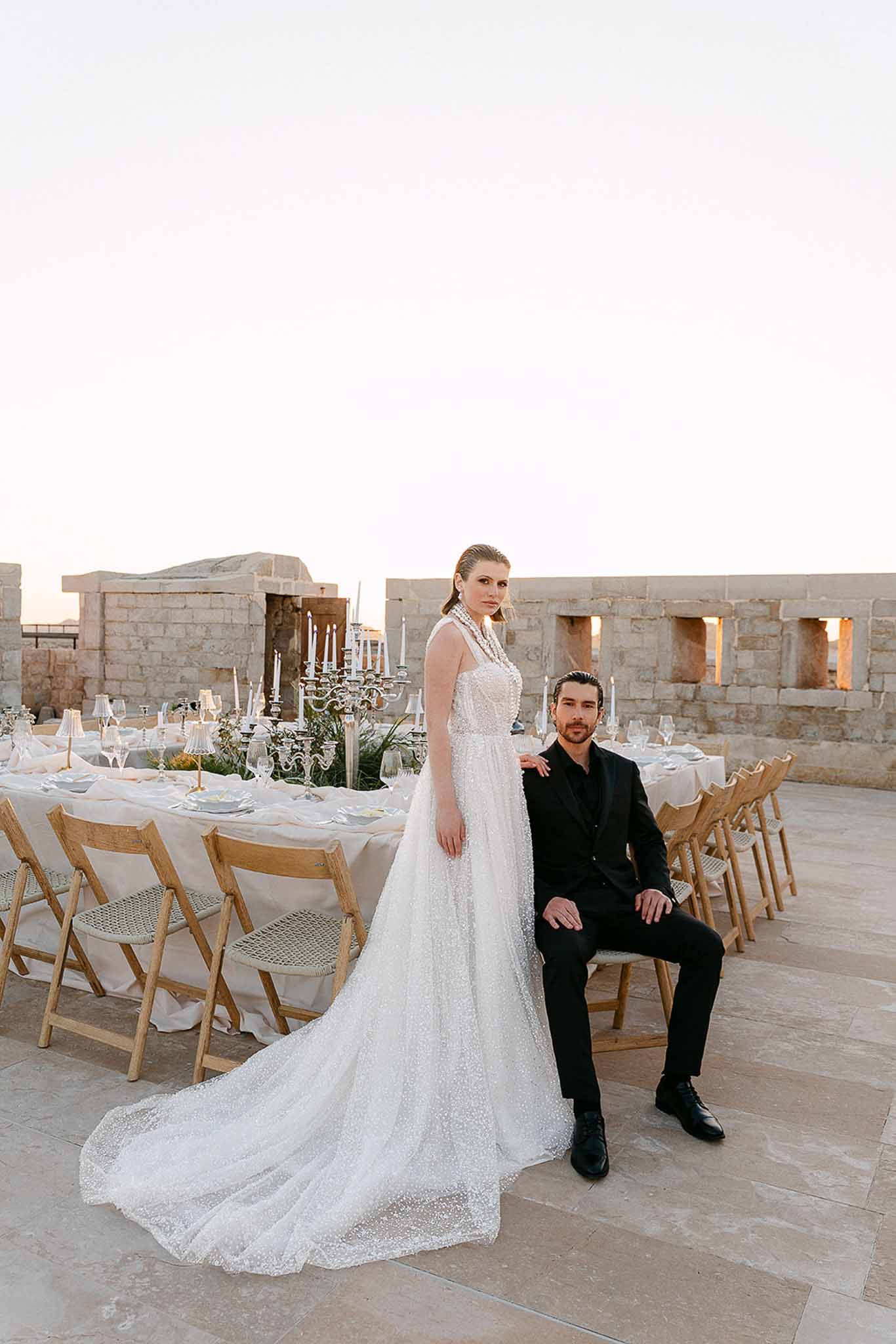 Bride in embellished gown and groom in black suit at stone terrace reception table with candelabras at dusk