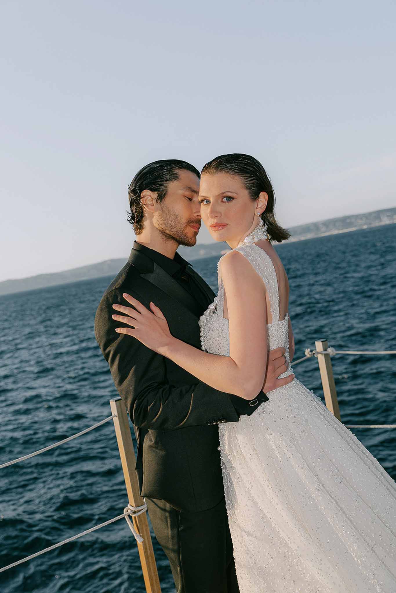 Couple embracing on sailboat stern with bride in beaded halter gown and groom in black mandarin-collar tux