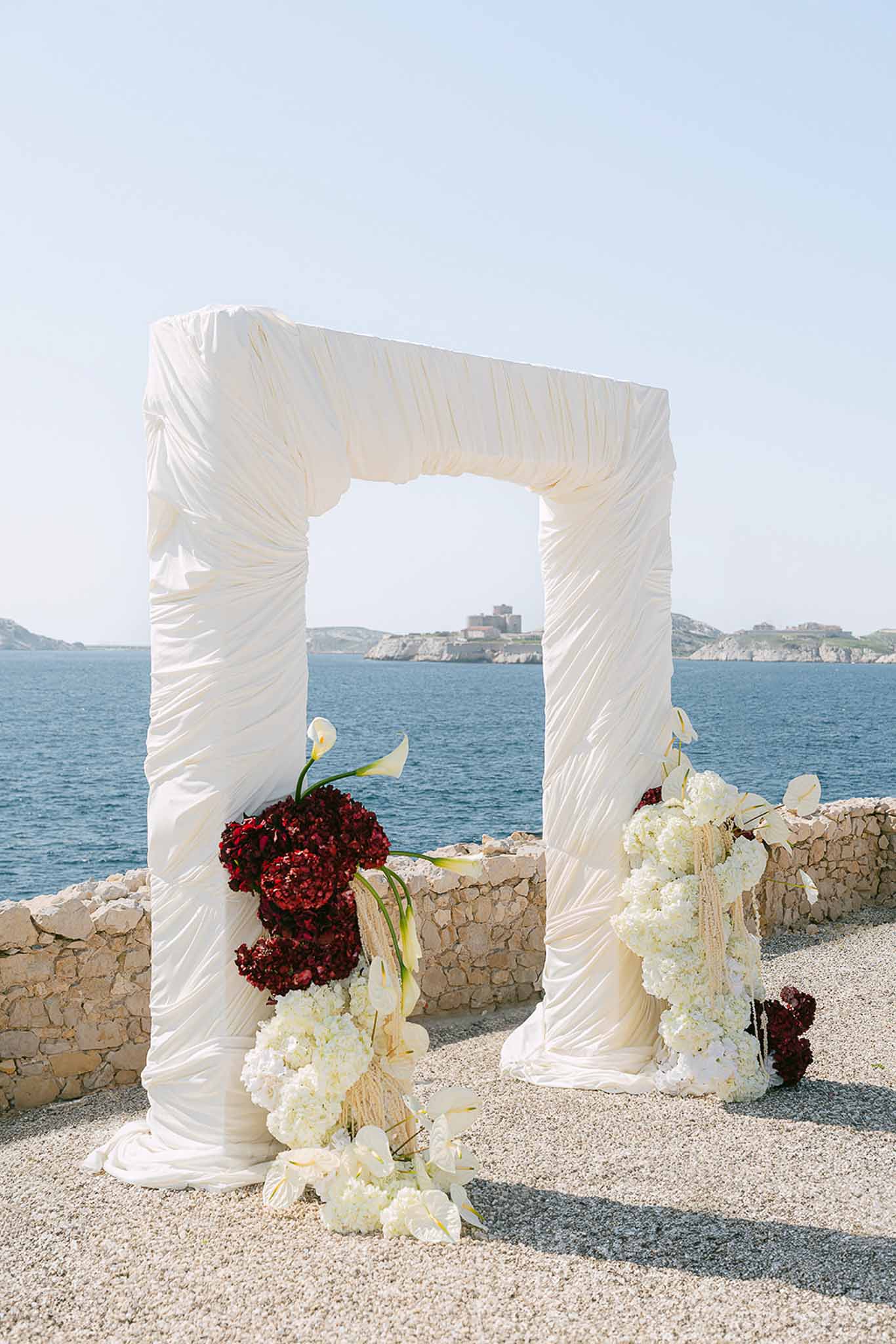 Draped ivory ceremony arch with burgundy and white floral arrangements on coastal terrace overlooking sea