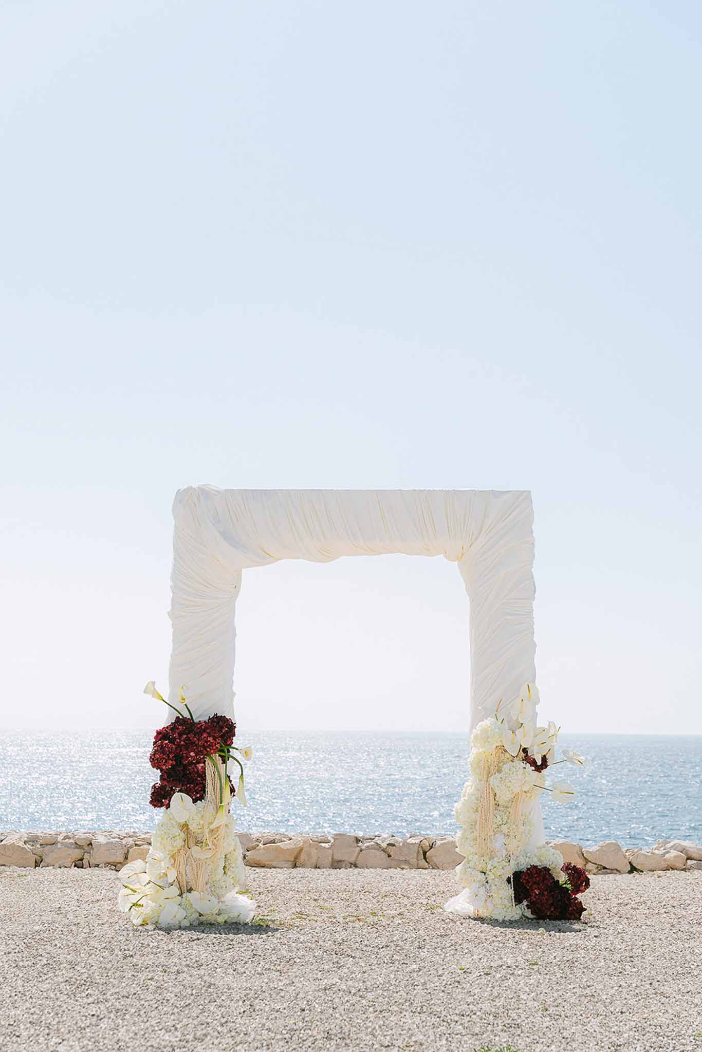 Fabric-draped ceremony arch with white hydrangeas and burgundy floral arrangements set against a seaside backdrop