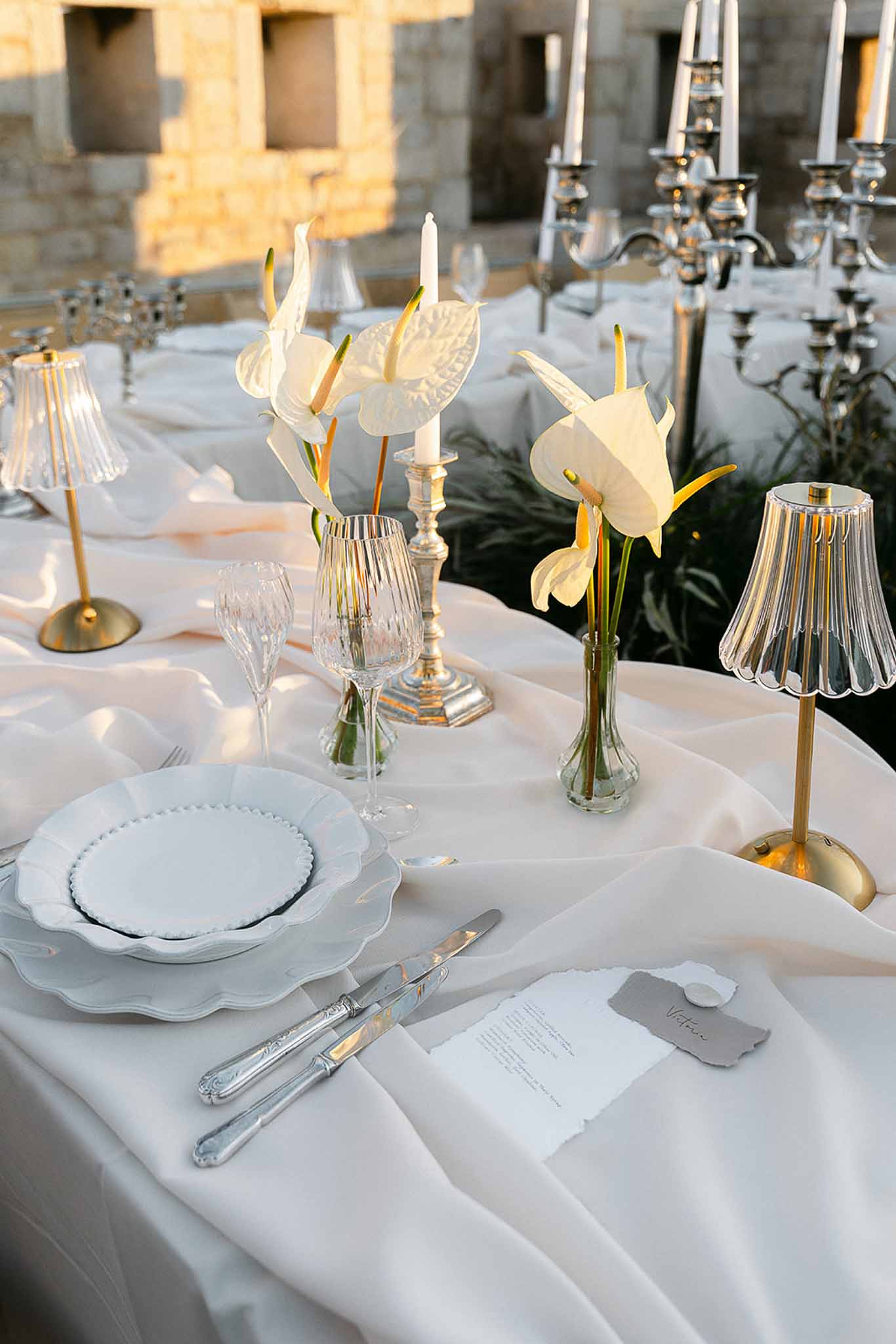 Golden-hour table setting with white anthurium bud vases, brass lamps, silver candelabra, and calligraphy cards